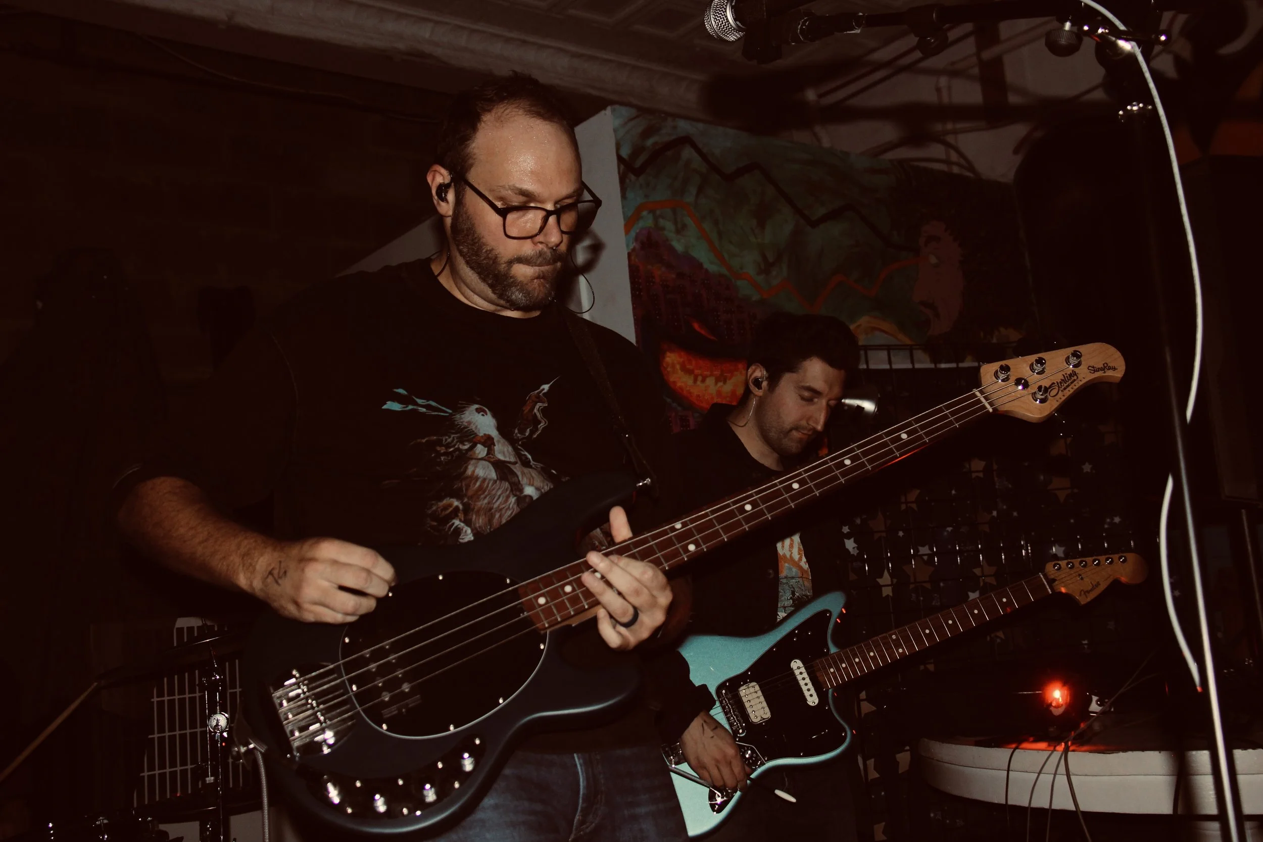 Two musicians performing with electric guitars in a dimly lit room. One musician is playing a black guitar, and the other is playing a light blue guitar. There is a colorful mural or artwork in the background.