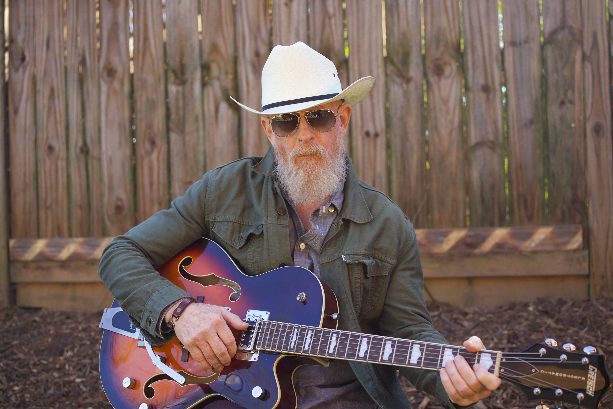 Man with white hat, sunglasses, and a beard playing an orange and blue electric guitar outdoors in front of a wooden fence.
