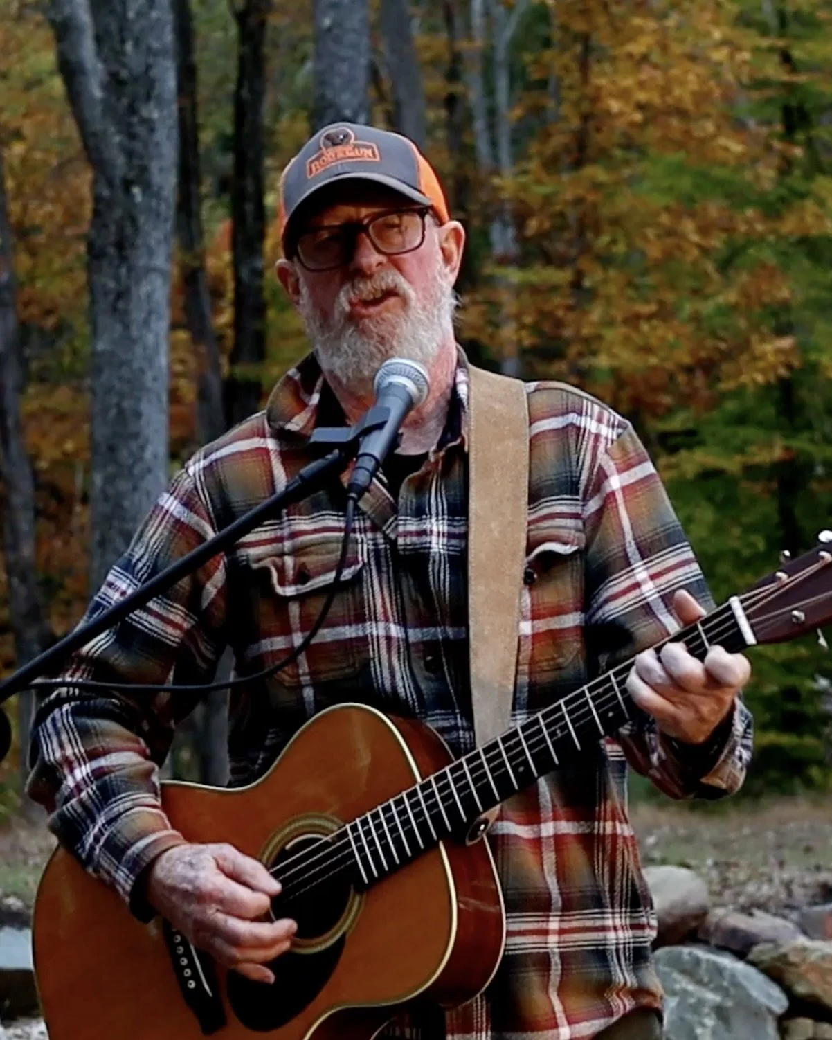 An elderly man with glasses, a beard, and wearing a cap, plaid shirt, and holding an acoustic guitar, is singing into a microphone outdoors in a wooded area with autumn foliage.