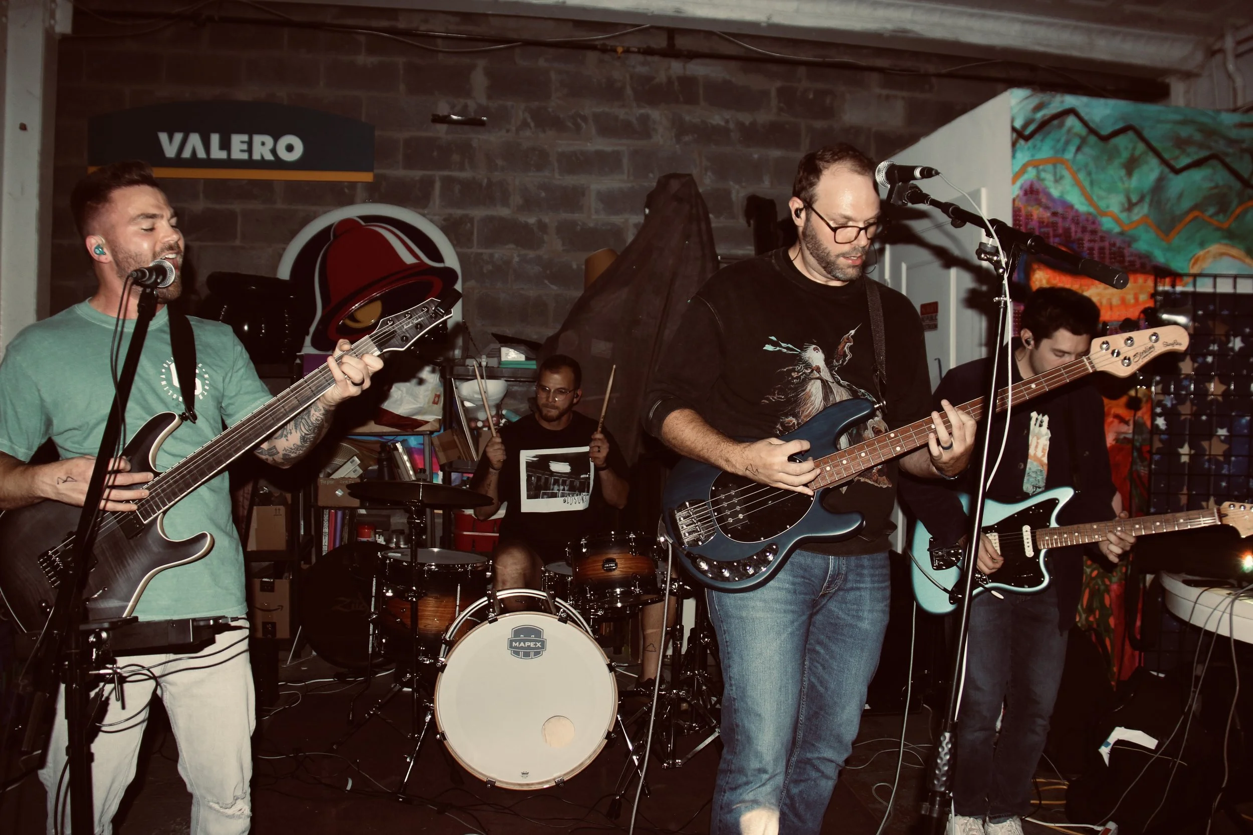 A band performing live music in a cozy indoor setting with brick walls, colorful artwork, and musical instruments, including guitars, drums, and microphones.