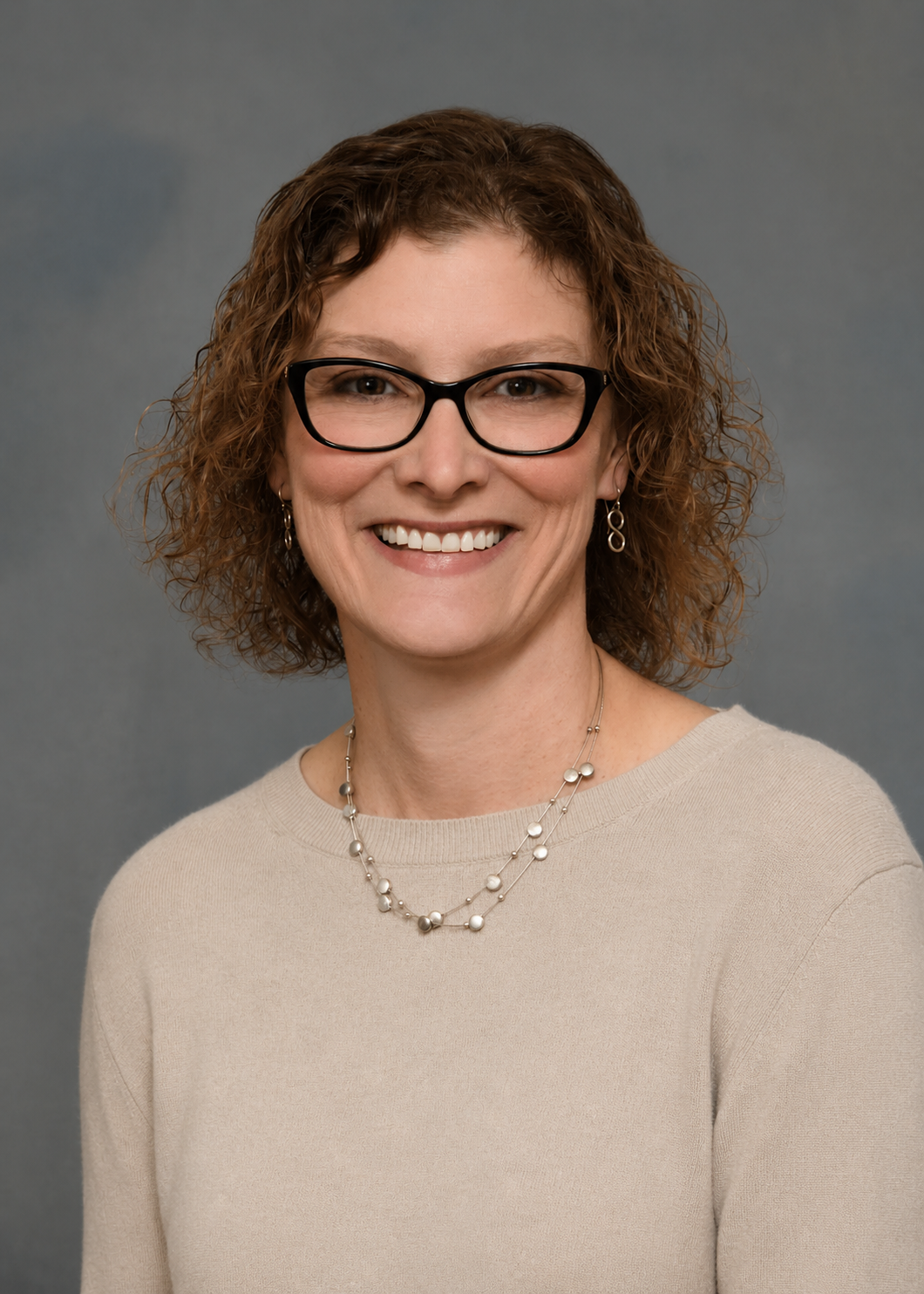 A woman with curly brown hair wearing glasses, pearl earrings, a necklace, and a beige top, smiling against a gray background.