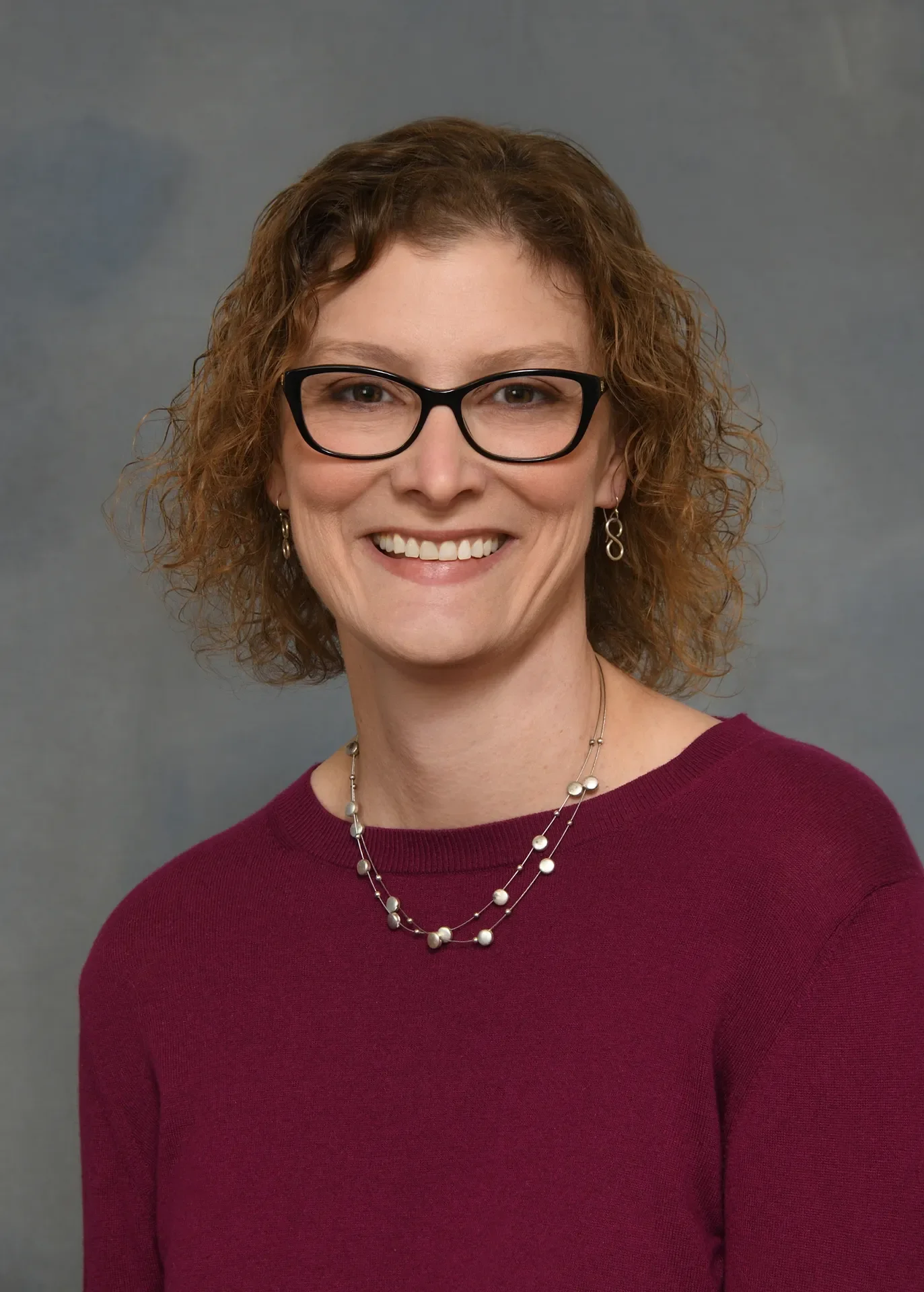 A woman with curly short brown hair and glasses, smiling, wearing a maroon top and a double-layer pearl necklace, standing against a gray background.