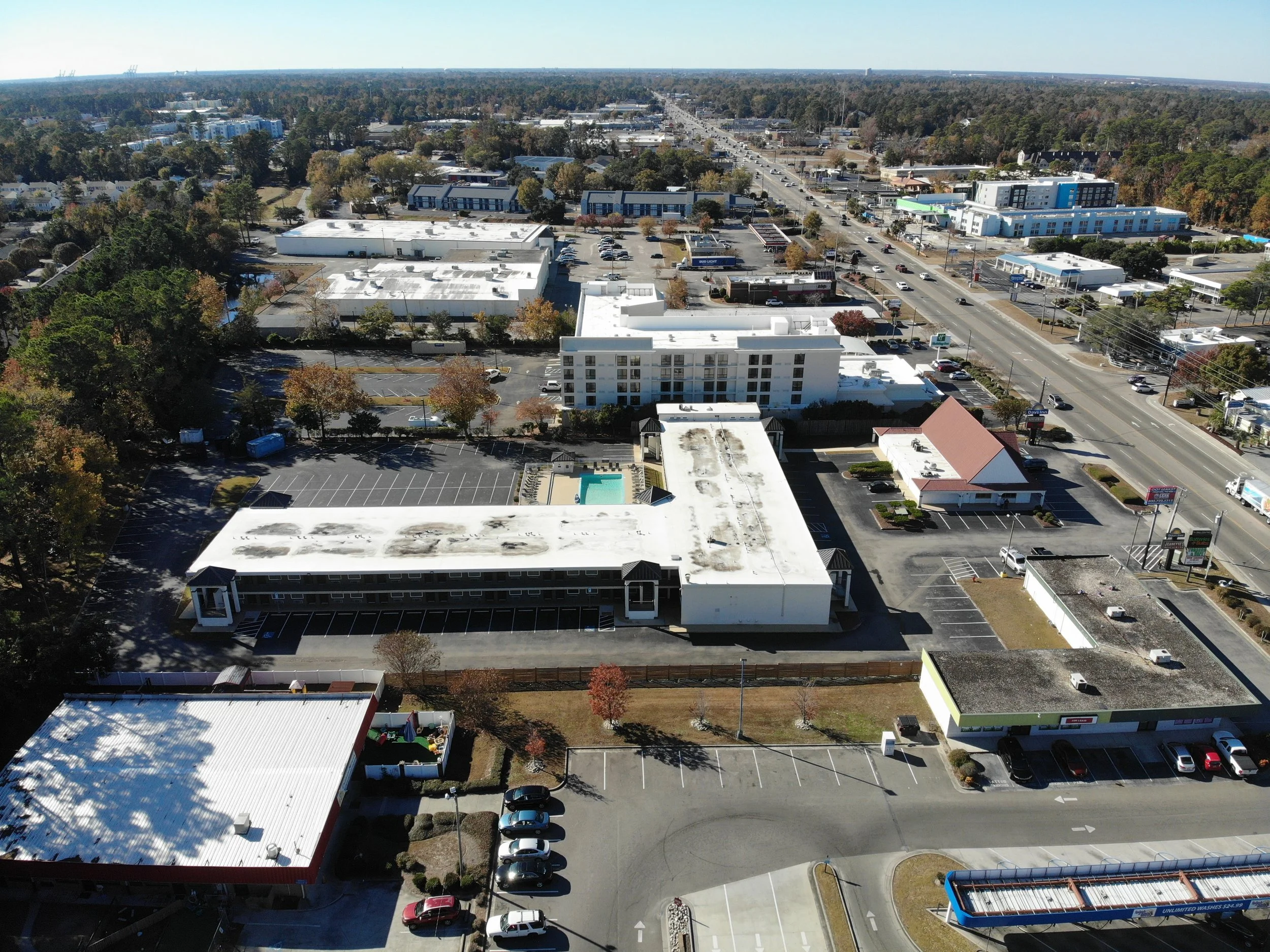 Aerial view of a commercial area with parking lots, buildings, and a busy road, with trees and distant cityscape.