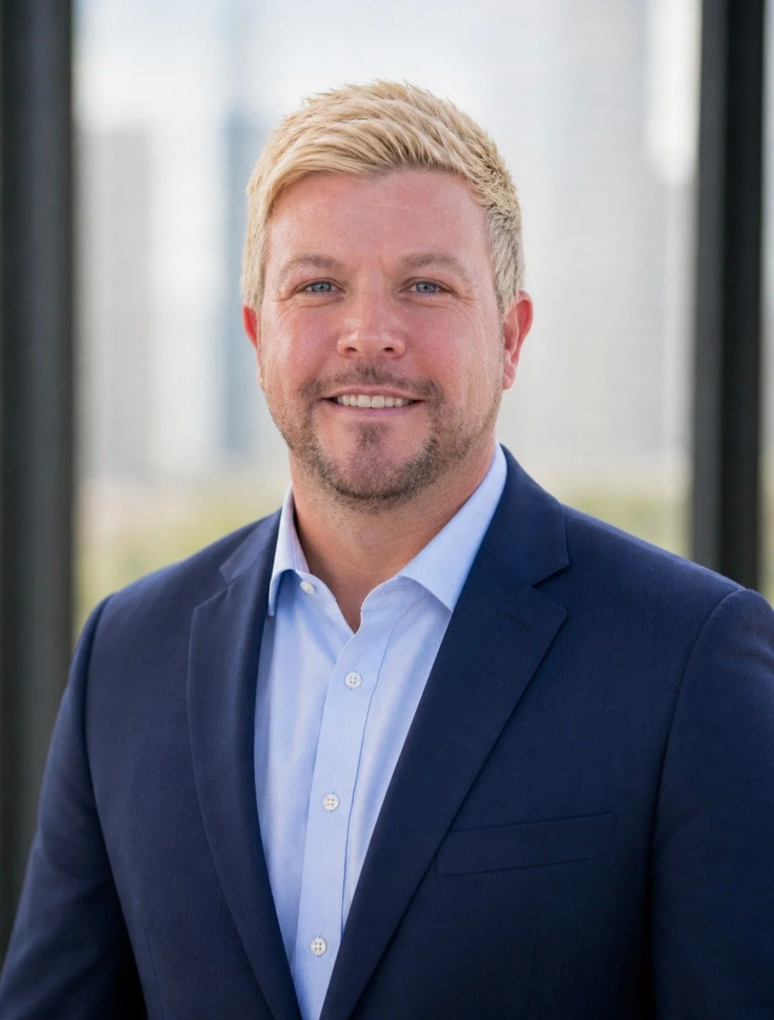 Portrait of a man with blonde hair, blue eyes, and a beard, wearing a navy suit and light blue shirt, standing indoors with a blurred cityscape background.