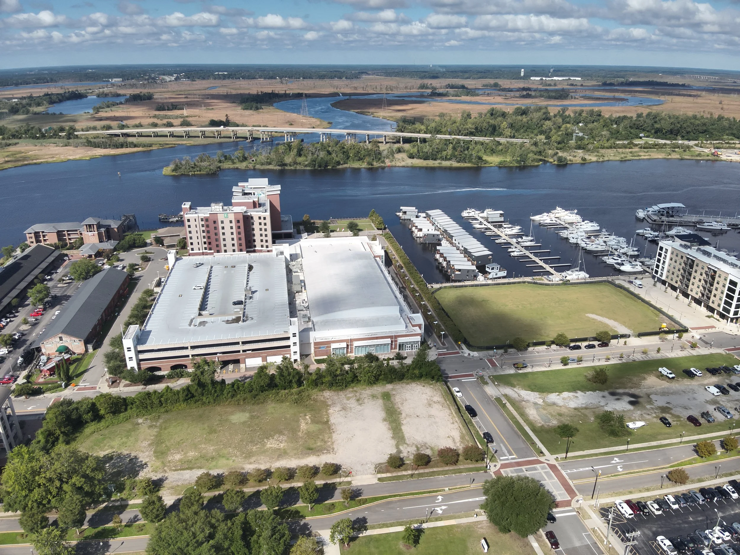 Aerial view of a city waterfront with a marina full of boats, a large parking lot, high-rise buildings, a river with bridges, and vast open land and rivers in the background under partly cloudy skies.