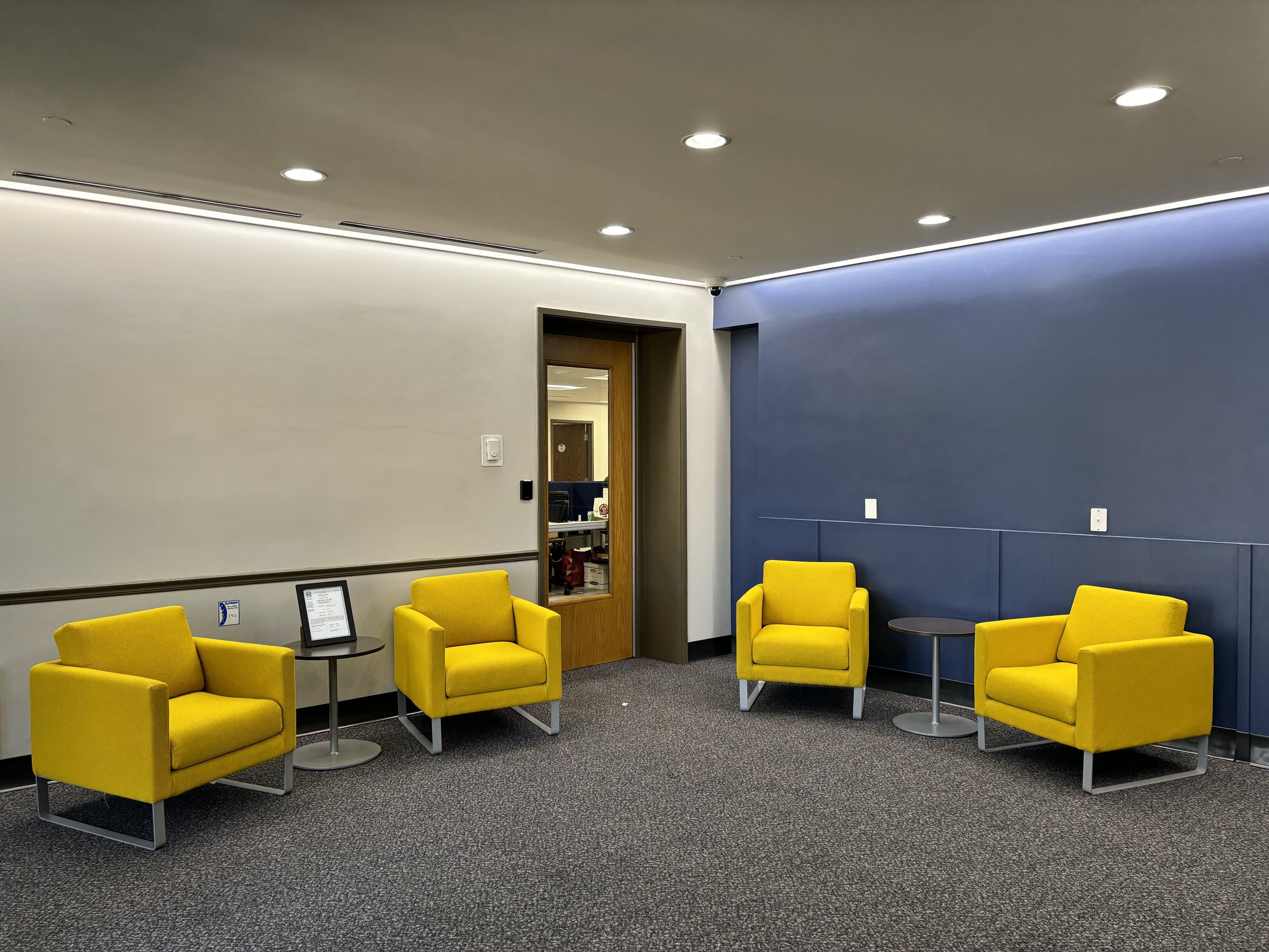 Corporate seating area with four yellow armchairs, small tables, a blue accent wall, and perimeter lighting.