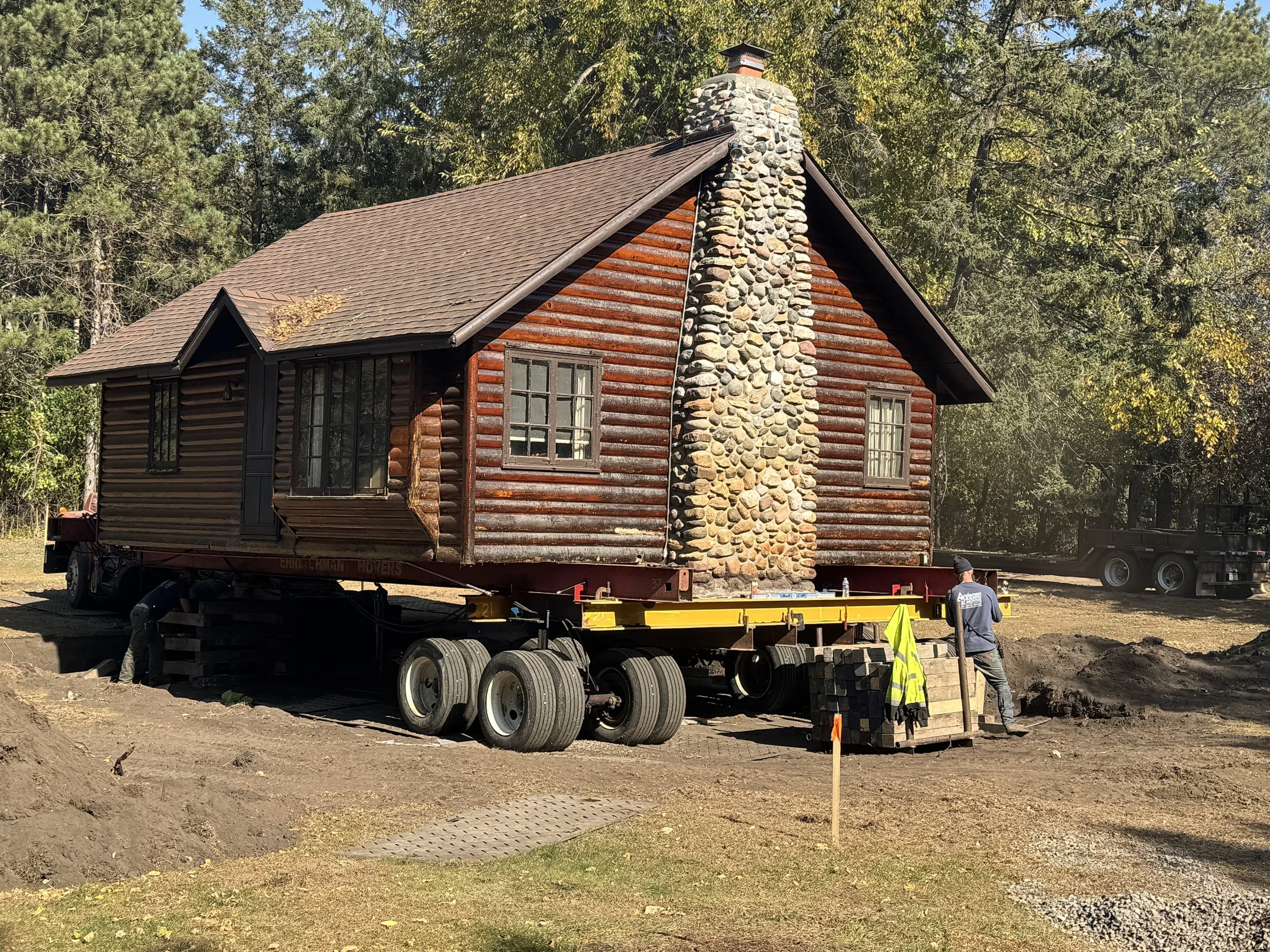 Historic log cabin supported on moving equipment during relocation to a new foundation and site.