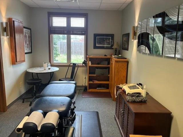 Chiropractic clinic treatment room with adjustment table, storage, seating, and natural light.