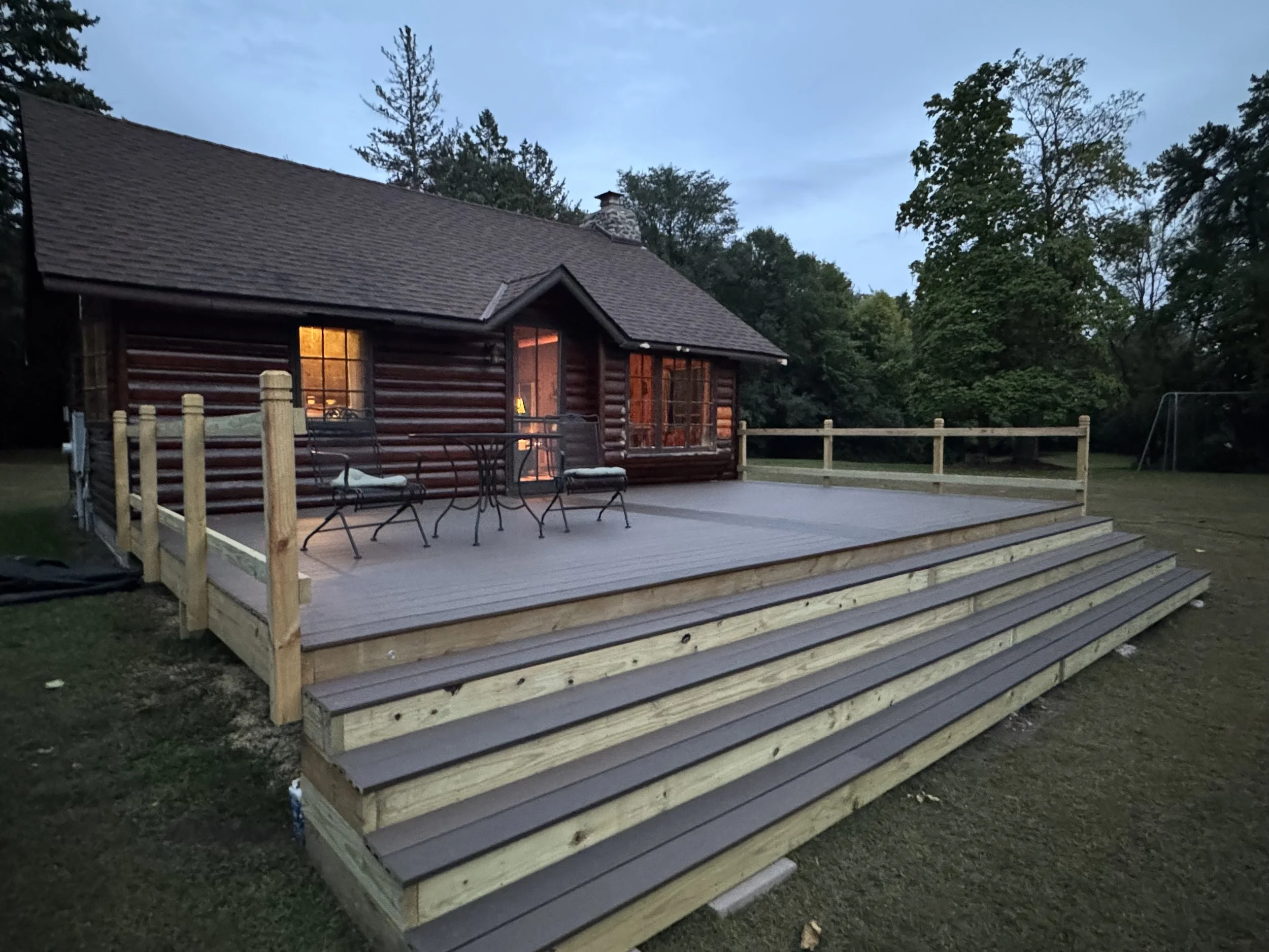 Historic log cabin at its relocated site with outdoor deck, surrounding trees, and yard.
