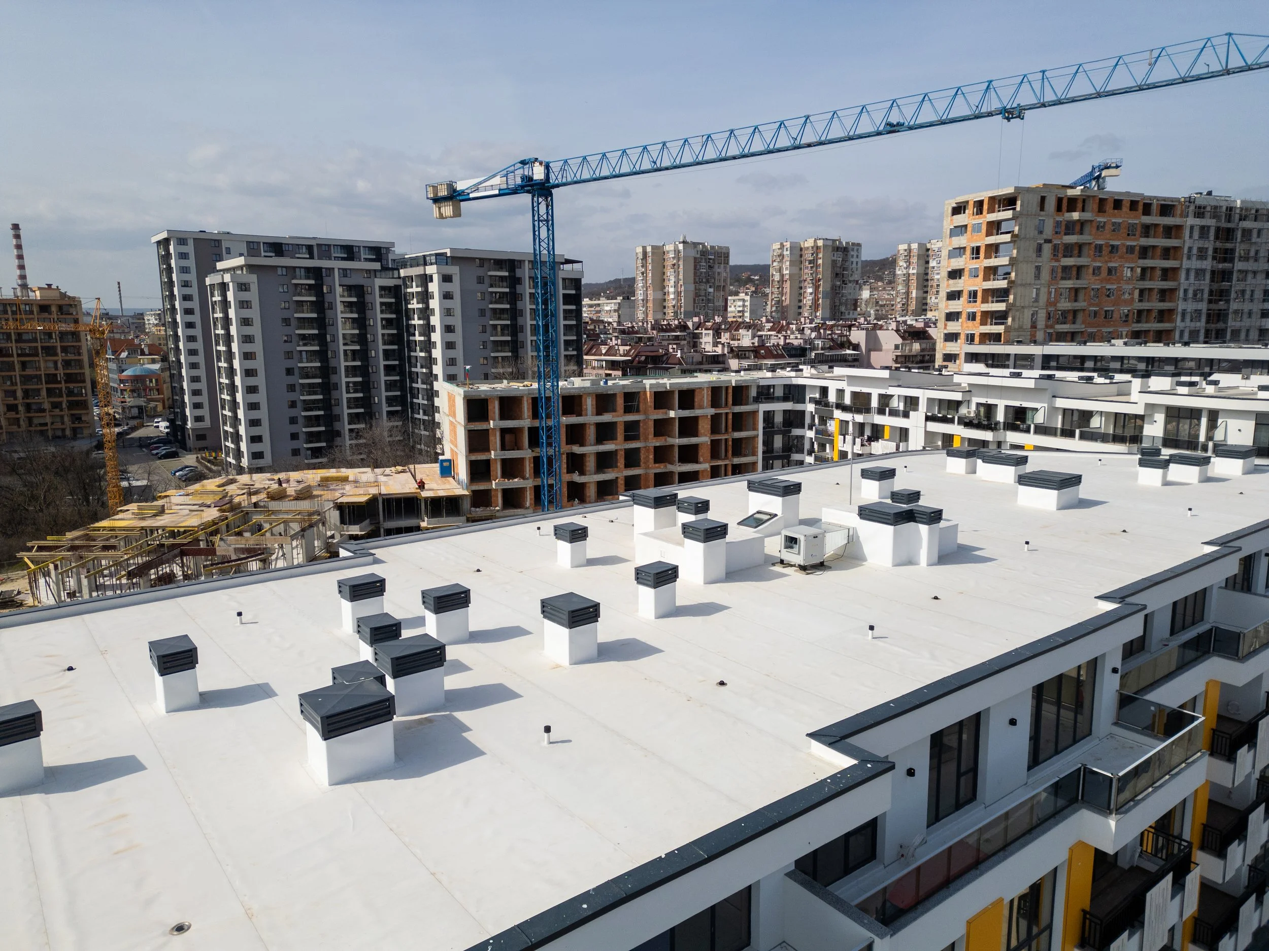 A white rooftop with multiple black vent pipes, overlooking a city with various modern buildings and a construction crane in the background.
