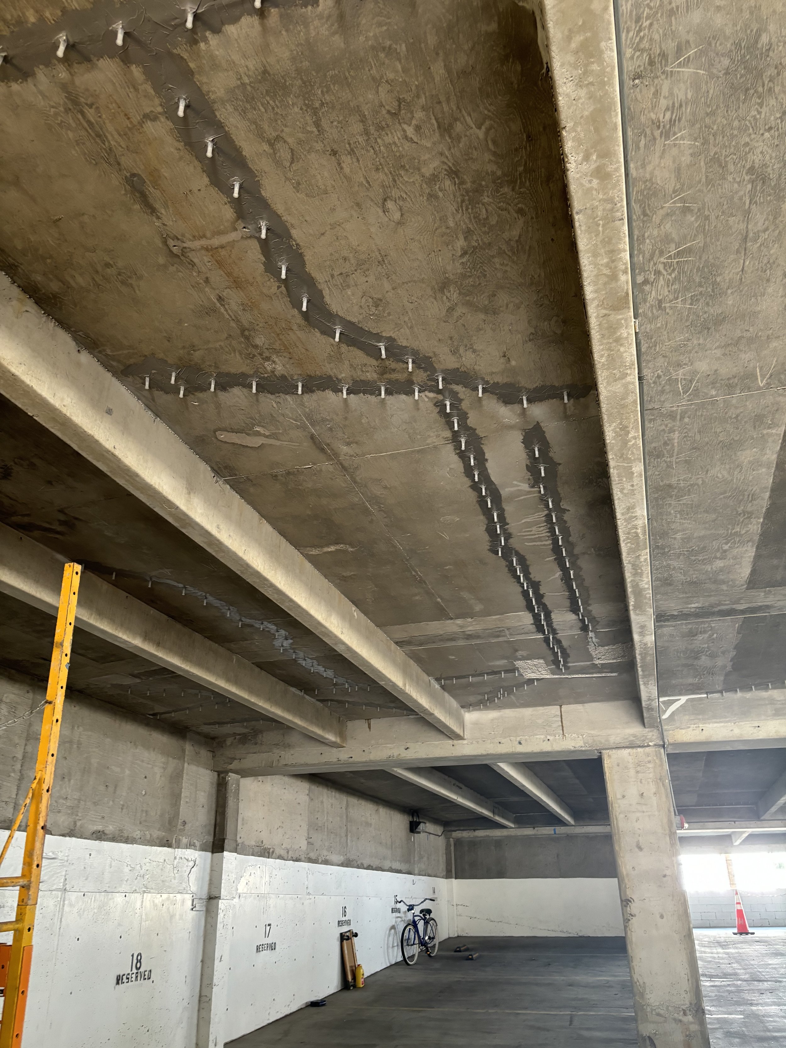 Underground parking garage under construction with unfinished concrete ceiling, concrete columns, bicycle, and reserved parking spots marked on the wall.