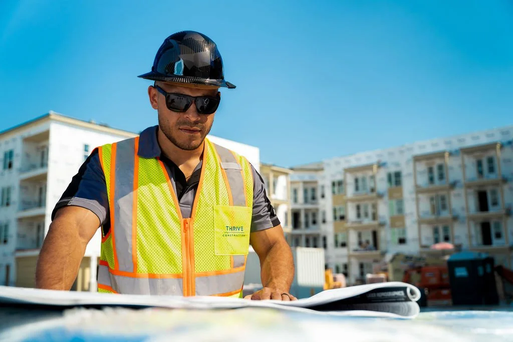 Construction worker wearing a safety vest and helmet reviewing blueprints at a building site with apartment buildings in the background on a sunny day.