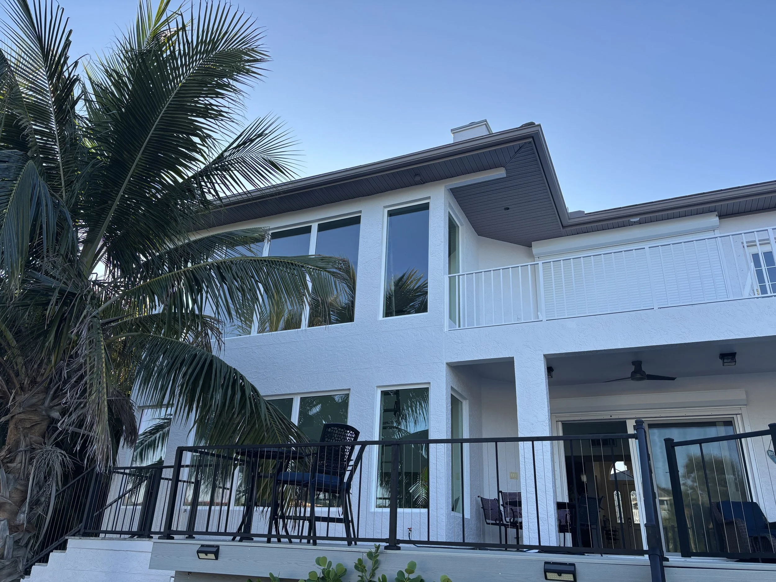 A modern white multi-story house with large windows, a balcony, and a small patio area. There is a palm tree in the foreground and a clear blue sky in the background.