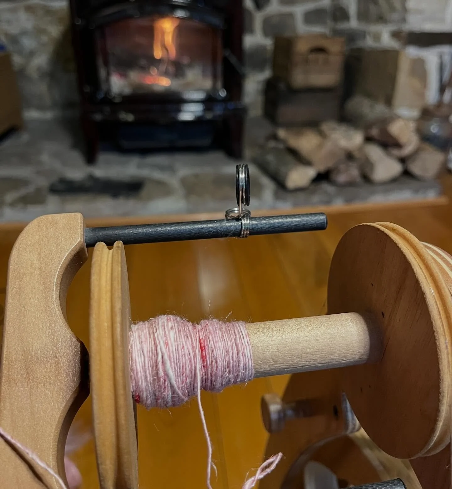 A close-up of a wooden spinning wheel with pink yarn, with a fireplace and stone hearth in the background.