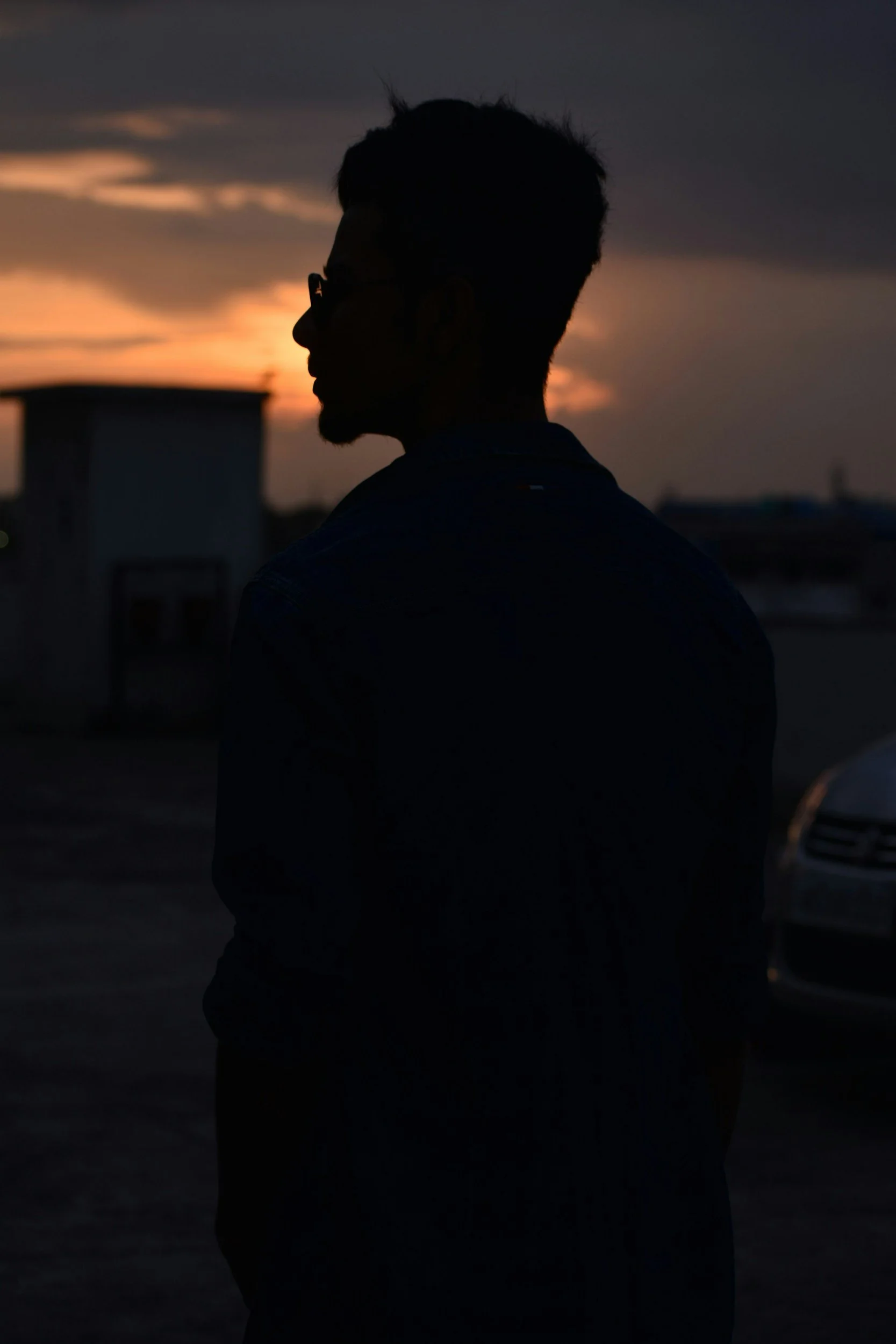 Silhouette of a person wearing glasses, standing outdoors during sunset or dusk, with a dark sky and blurred background.
