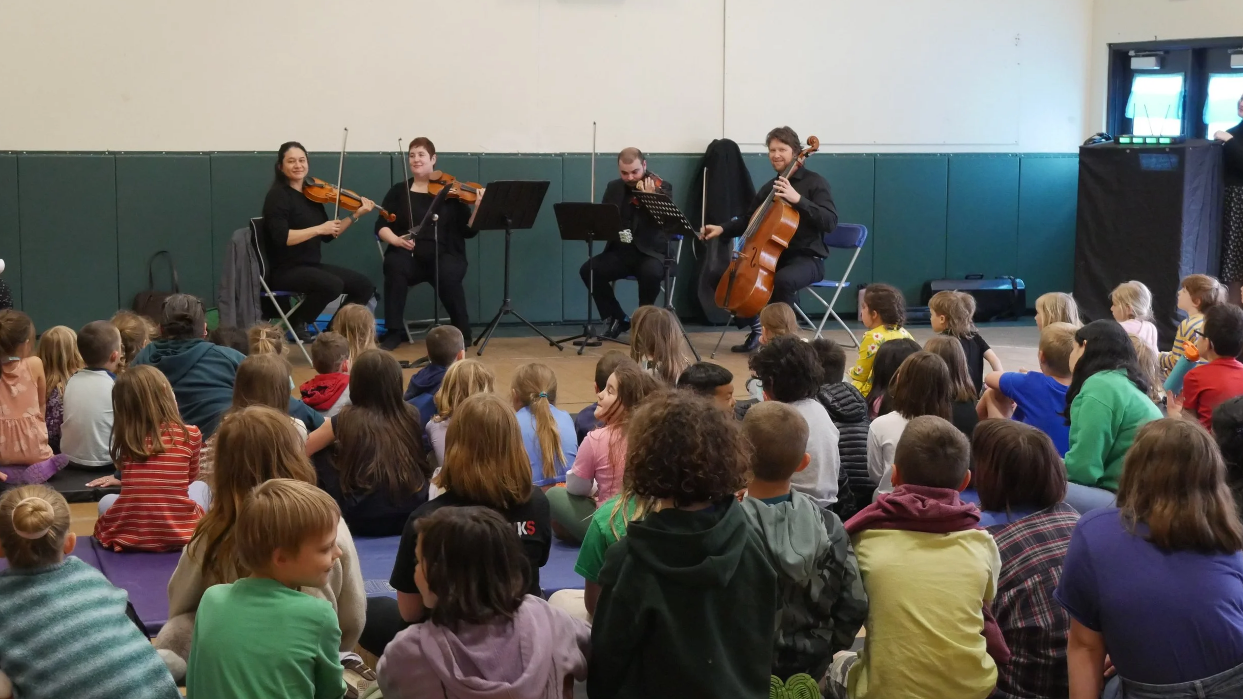 A group of four musicians performing for a large audience of children seated on the floor in a gymnasium or community center. The musicians are playing string instruments: two violins, a viola, and a cello.