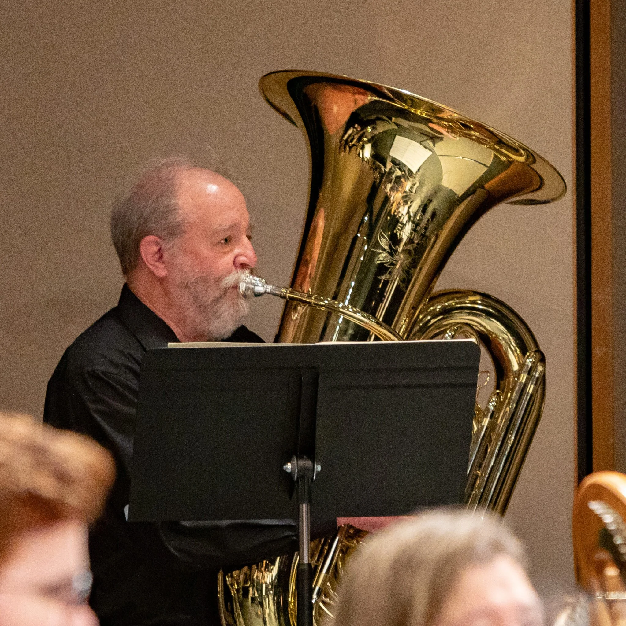 A man playing a brass tuba, viewed from the side, with part of his face and head visible, wearing a dark shirt and seated at a piano.