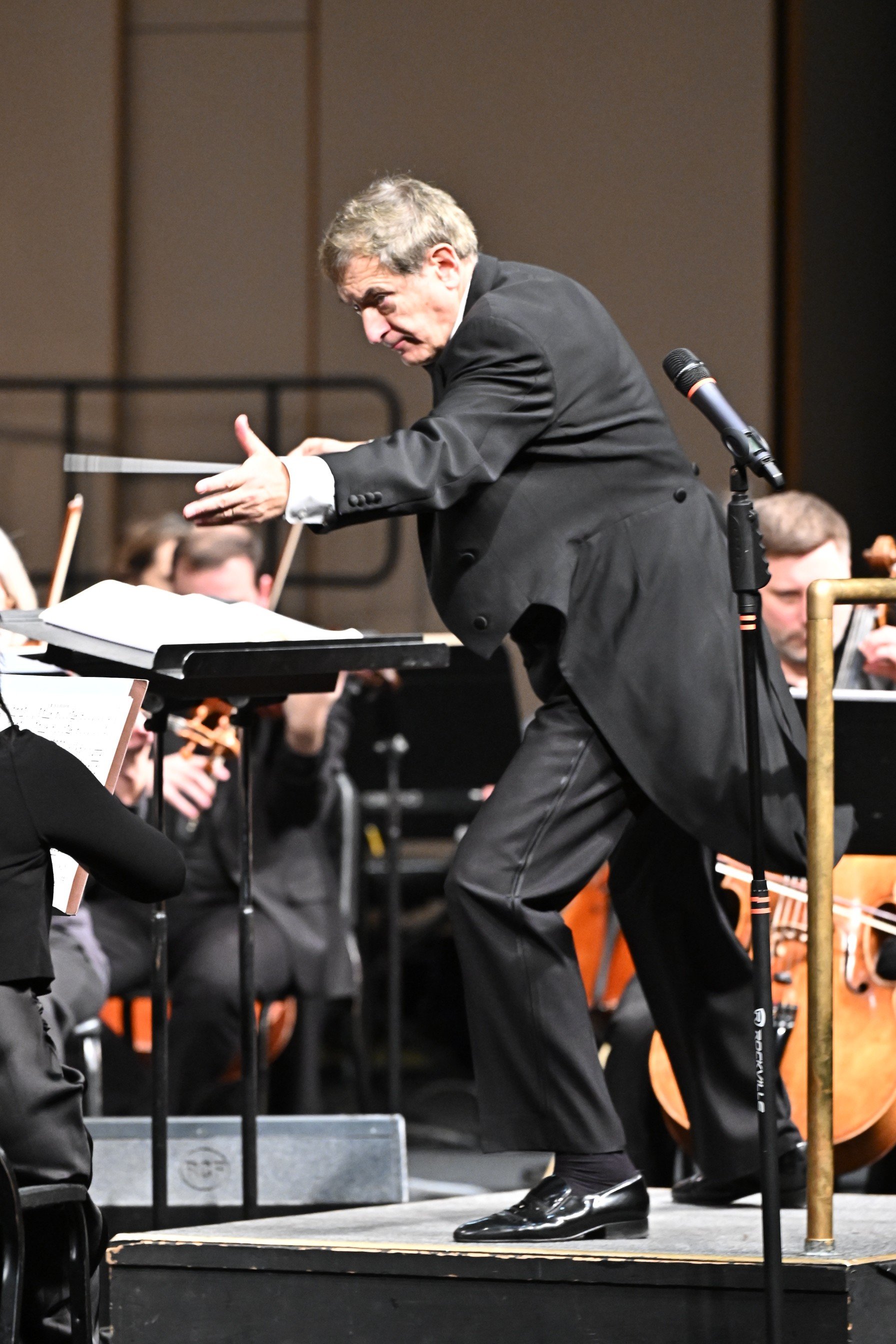 A conductor dressed in a black suit leading an orchestra, standing on a stage with a music stand and a microphone, with musicians playing string instruments in the background.