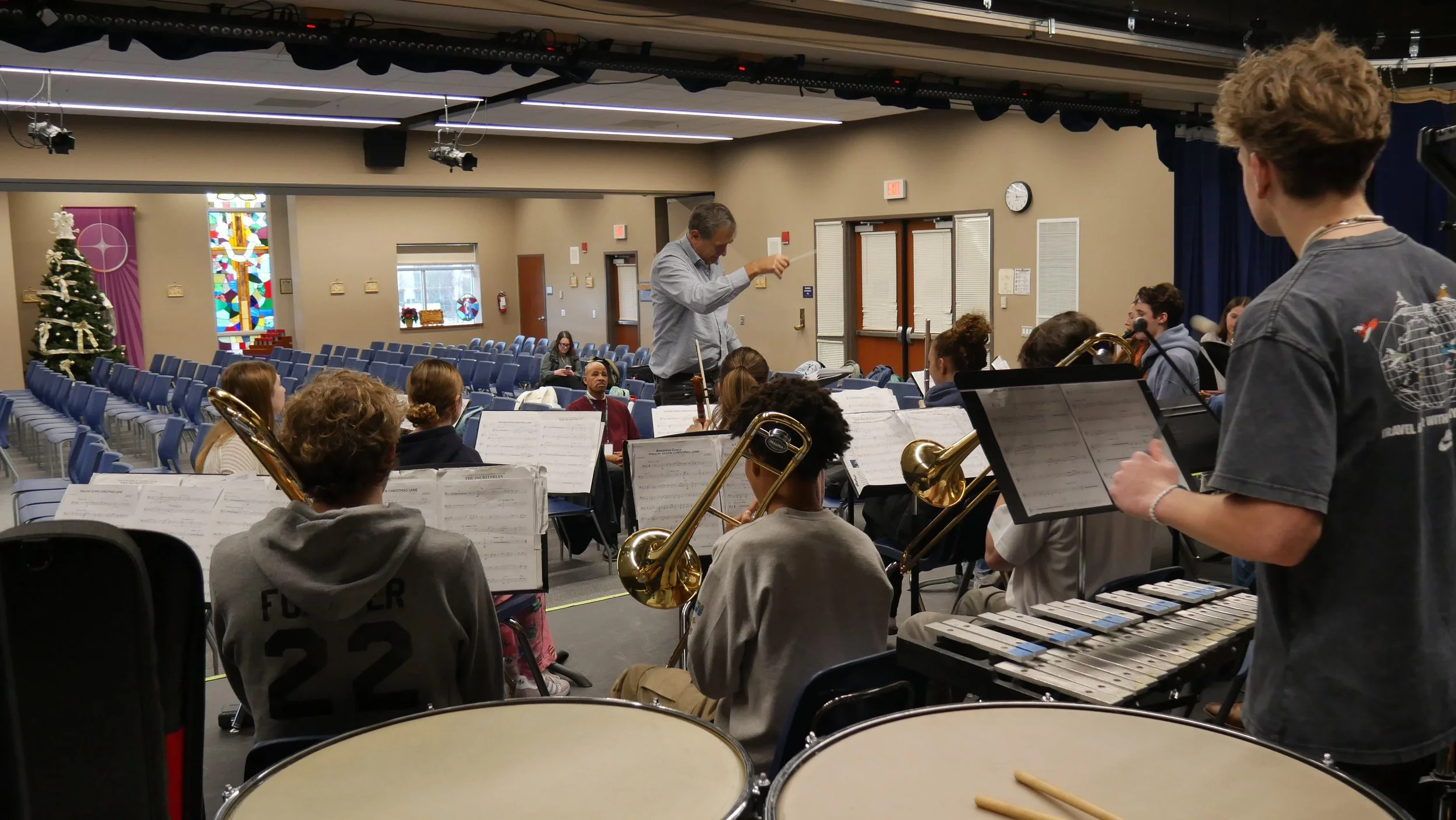 A group of young musicians practicing in a church or concert hall, with a conductor leading them. There is a decorated Christmas tree and stained glass windows in the background.