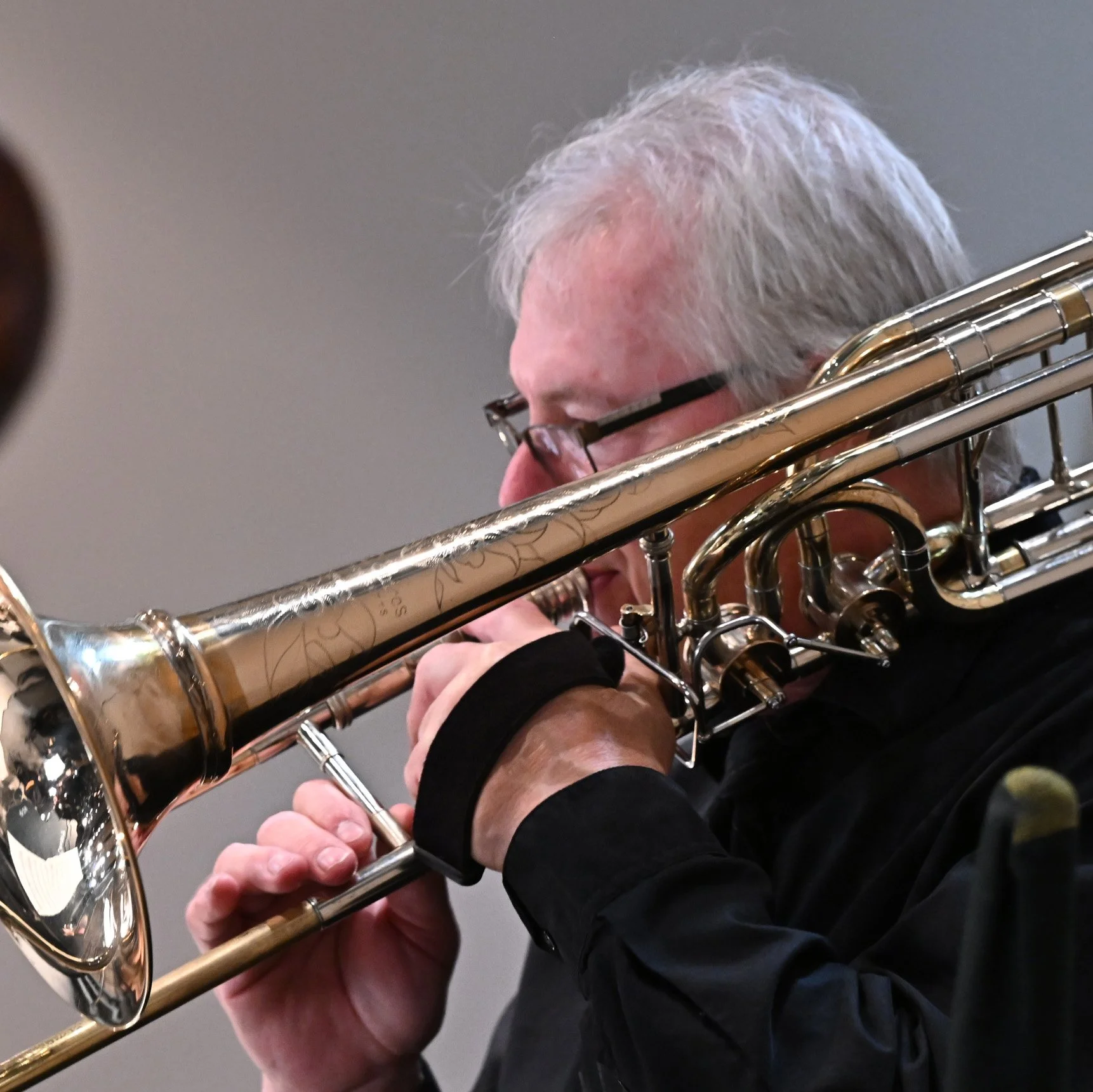 Close-up of a person playing a brass flute, focusing on the instrument and hands, with a gray background.