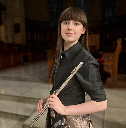 Young woman standing indoors holding a silver flute in her right hand, wearing a black blouse with puffy sleeves, with a wooden bench in the background.