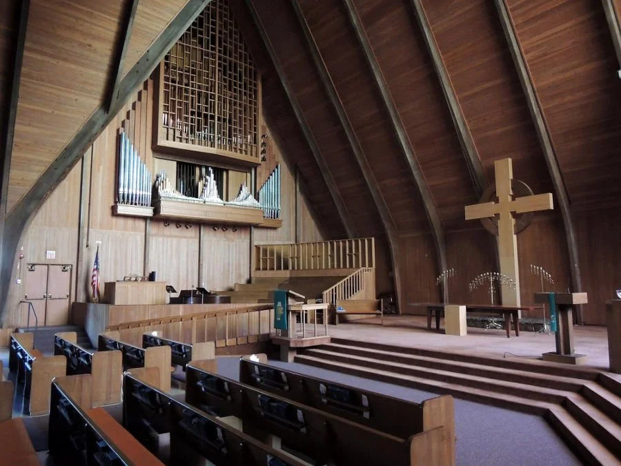Interior of a church with wooden pews, a large cross on the stage, and an organ at the back.