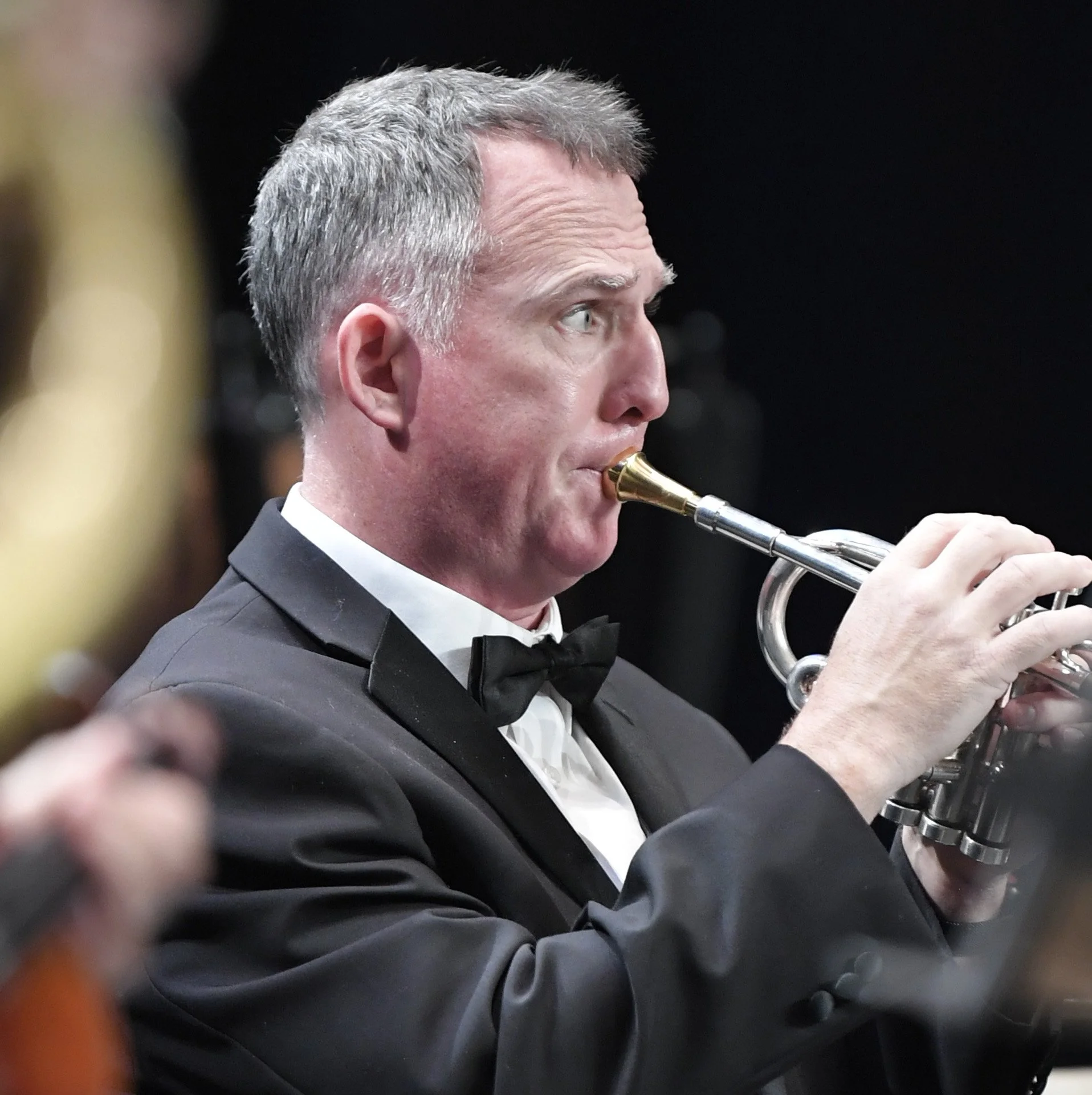 A man with short gray hair wearing a tuxedo and bow tie, playing a trumpet at a formal event.
