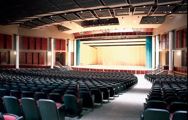 Empty auditorium with a large stage, rows of black chairs, and a high ceiling with lighting panels.