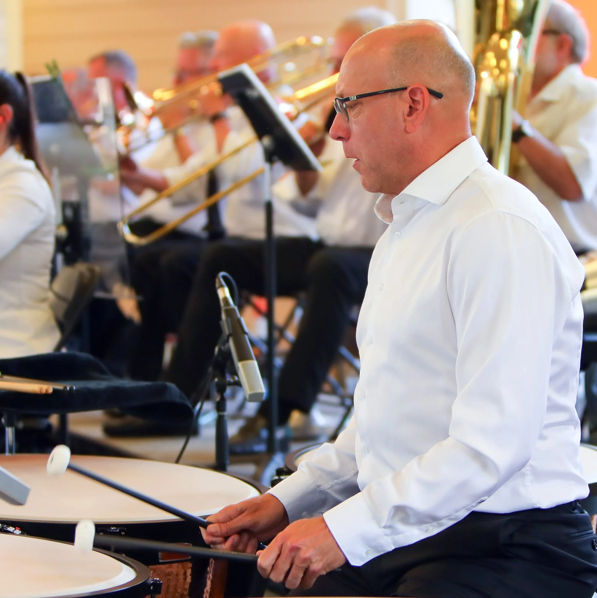 A man with glasses wearing a white shirt playing a keyboard or piano with a music stand nearby in a room with other musicians in the background.