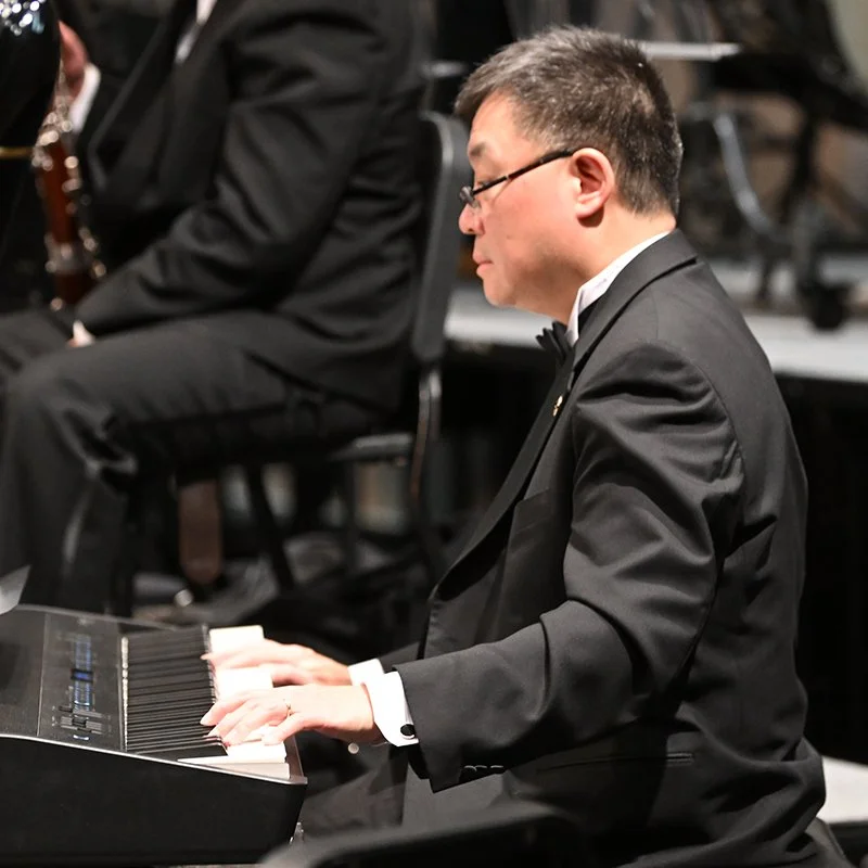 A man wearing glasses and a tuxedo playing the piano at an indoor concert.