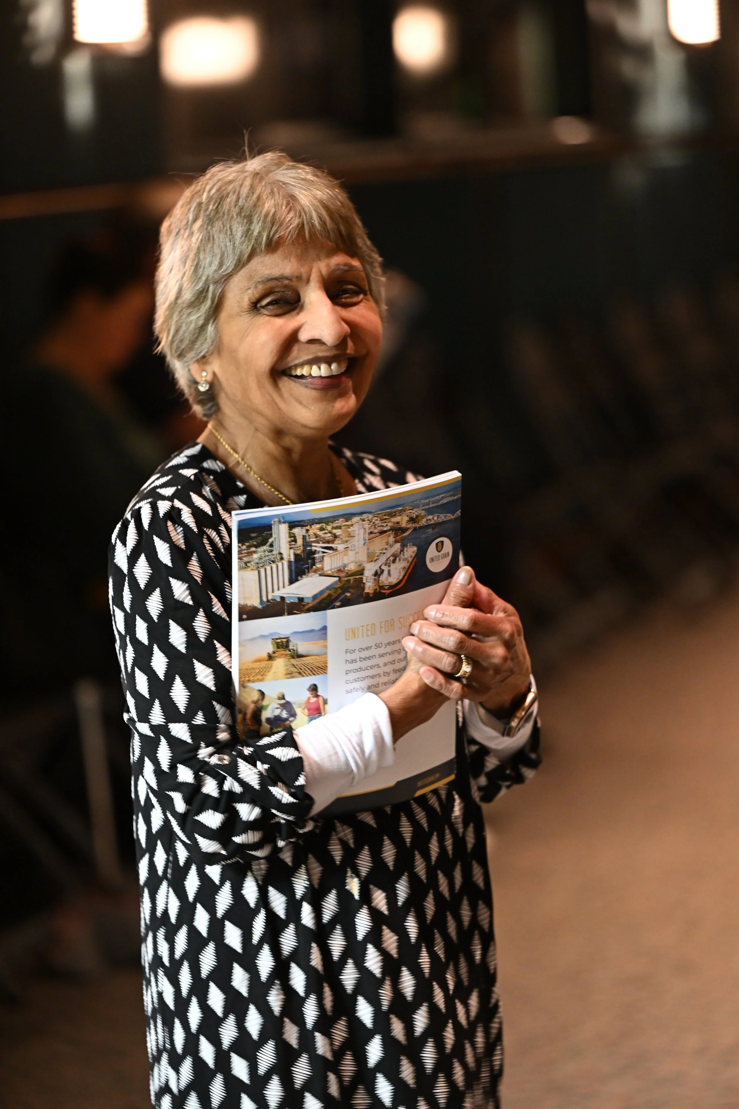 A woman with short gray hair smiling and holding a brochure, wearing a black and white patterned dress, standing indoors with blurred background lights.