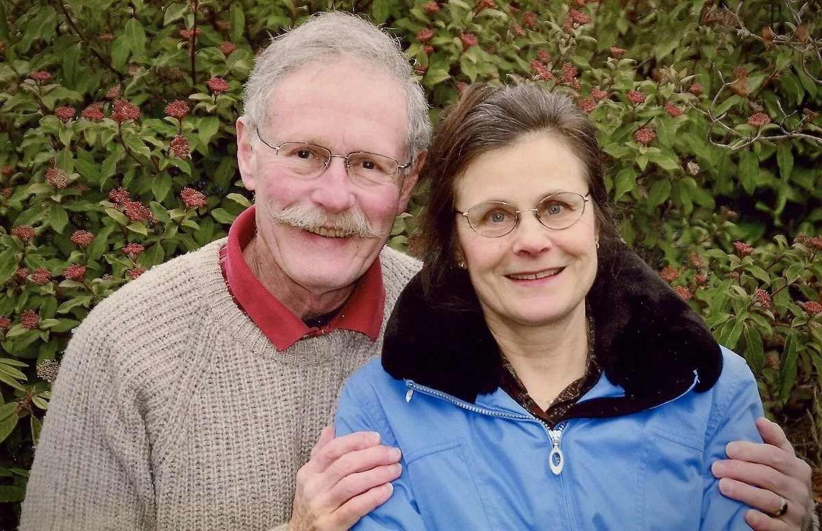 A smiling older man in glasses and a beige sweater standing next to a smiling woman with glasses and dark hair in a blue jacket with a black collar, outdoors in front of a bush with green leaves and red flowers.