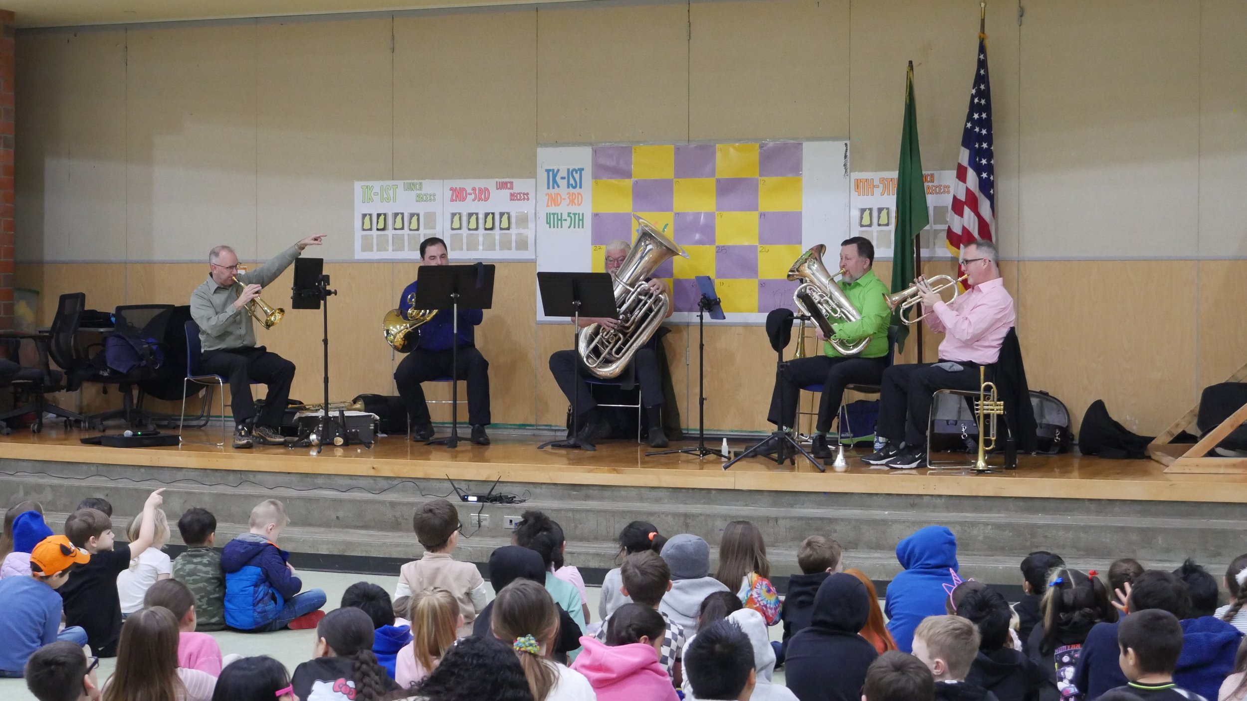 A group of five musicians playing wind instruments on stage in front of a school audience, with children sitting on the floor watching. The musicians are dressed in colorful shirts and are playing brass and woodwind instruments, with a background of a bulletin board, American flag, and green flag.