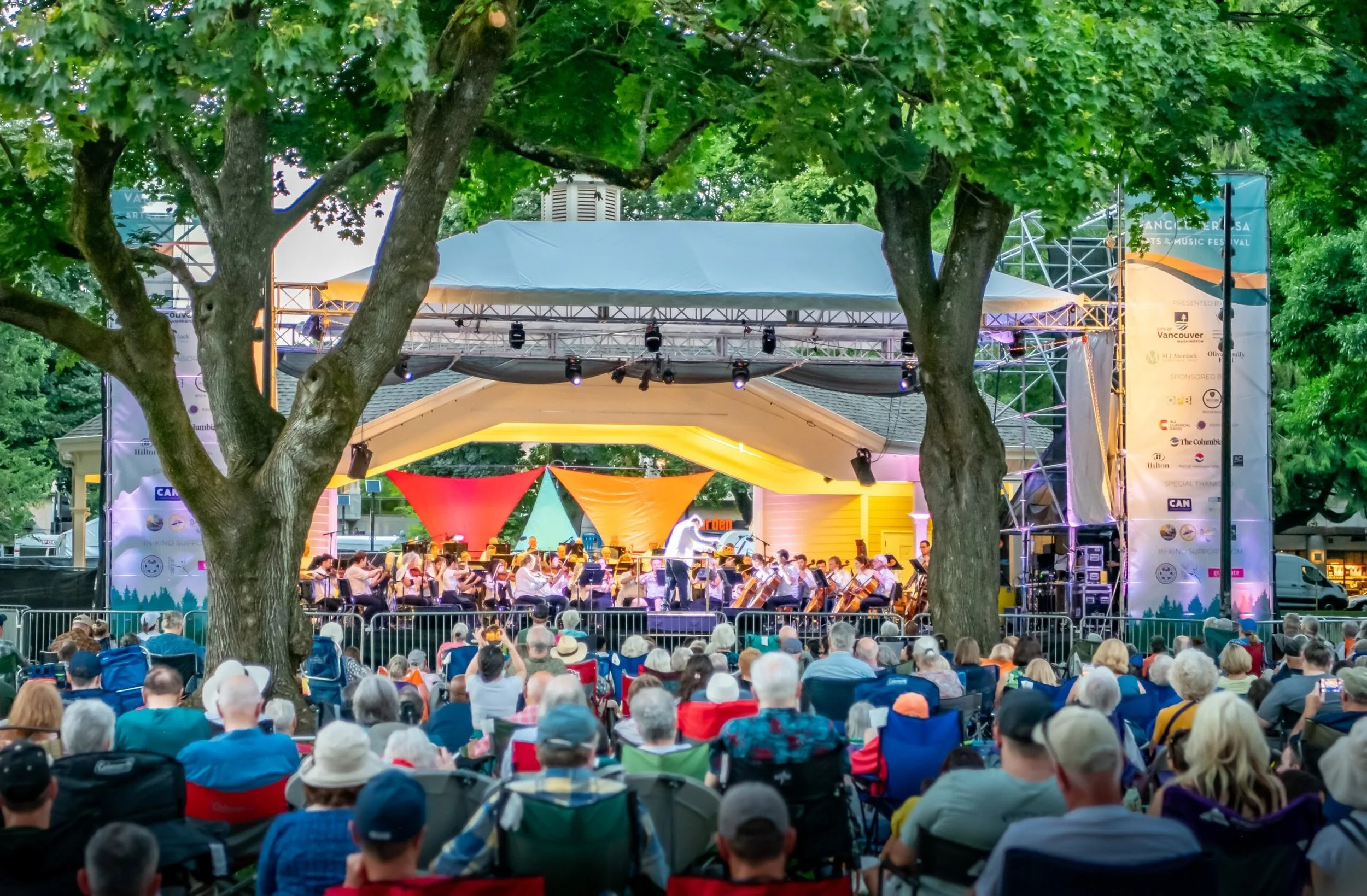 An outdoor concert stage with an orchestra playing, surrounded by many seated spectators. Trees frame the scene, and festival banners are visible on the sides of the stage.