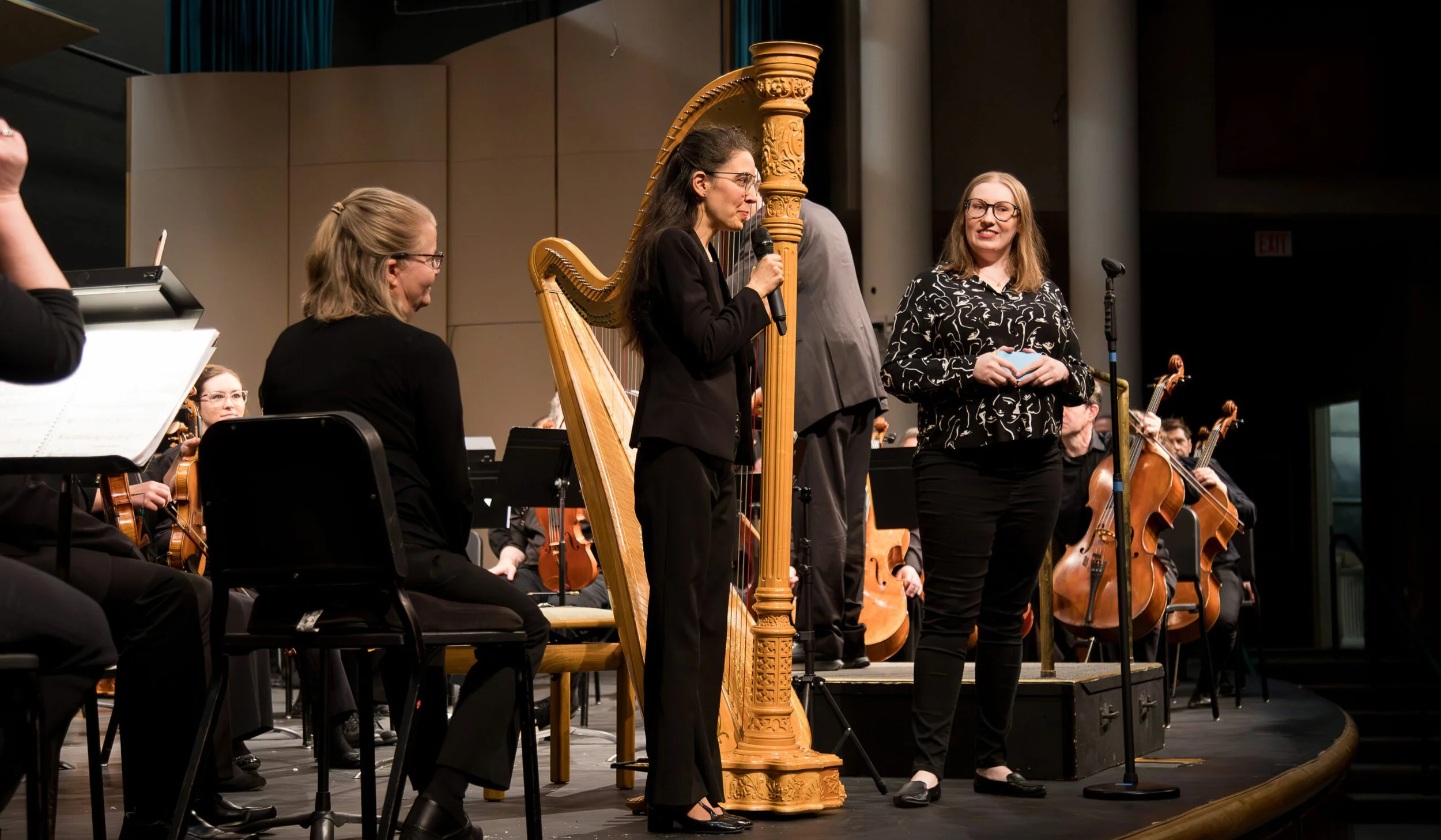 Orchestra performance with musicians playing violins and cellos, while two women speak on stage, one of whom is standing at a harp and the other holding a microphone.