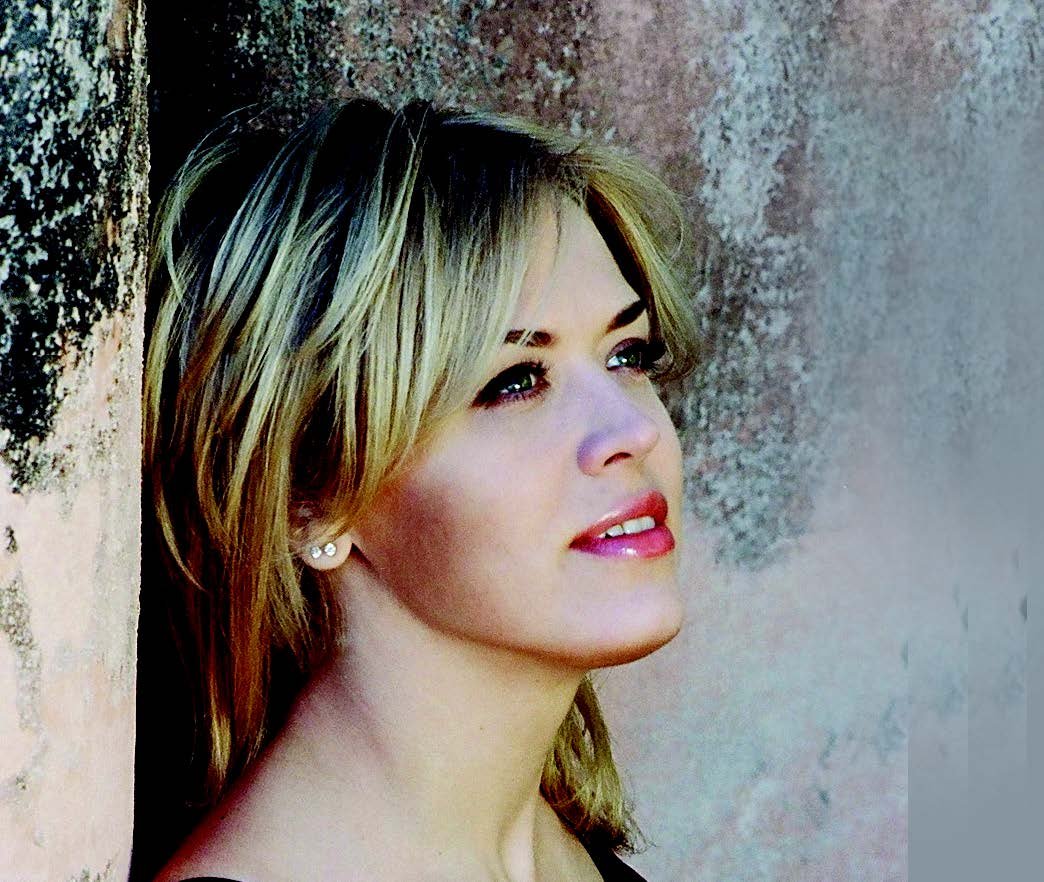 Close-up of a young woman with blonde hair and earrings, leaning against a textured wall, looking thoughtfully to the side.