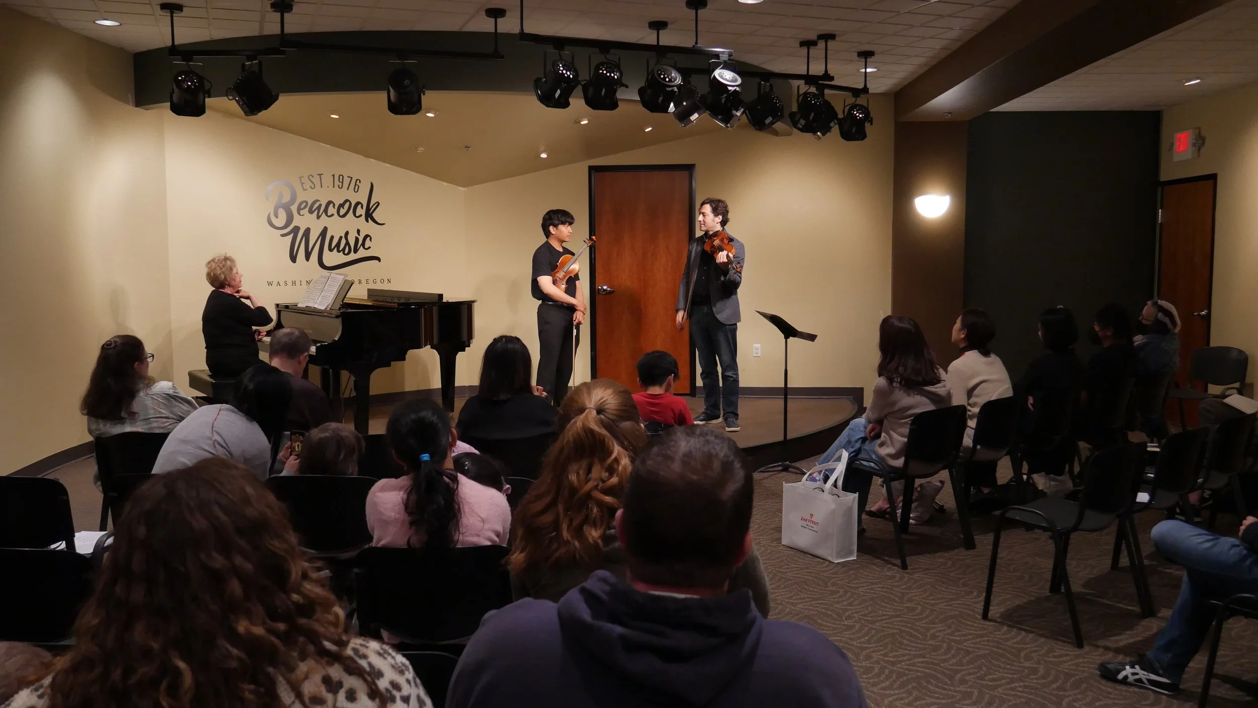 A performance on stage at Beacock Music in Washington, Oregon. Two musicians are playing violins while a woman at a piano accompanies them. Audience members are seated and watching attentively.
