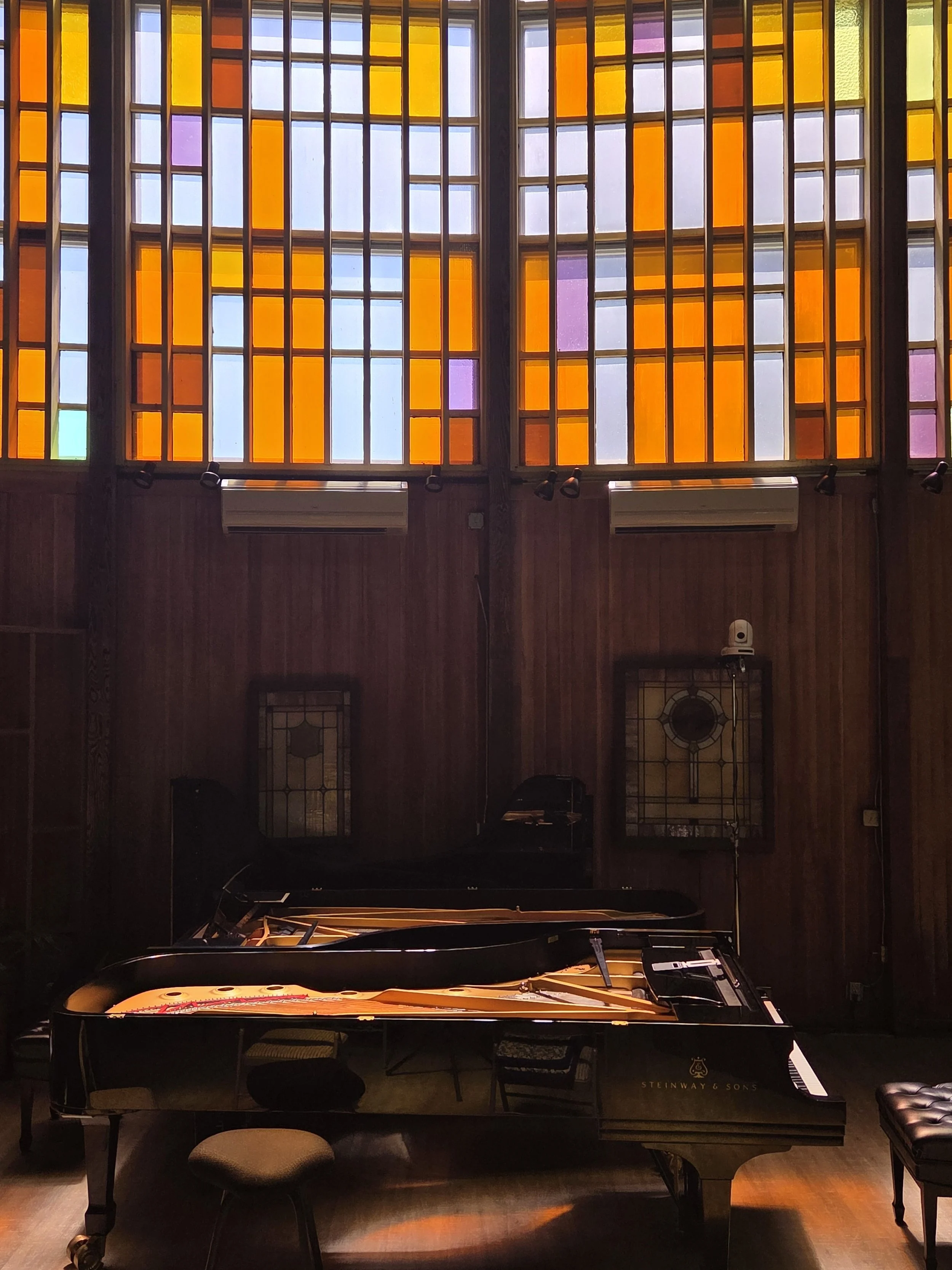 A grand piano with the brand name Steingray & Sons in a room with large stained glass windows made of colorful glass panes, illuminated by natural light.
