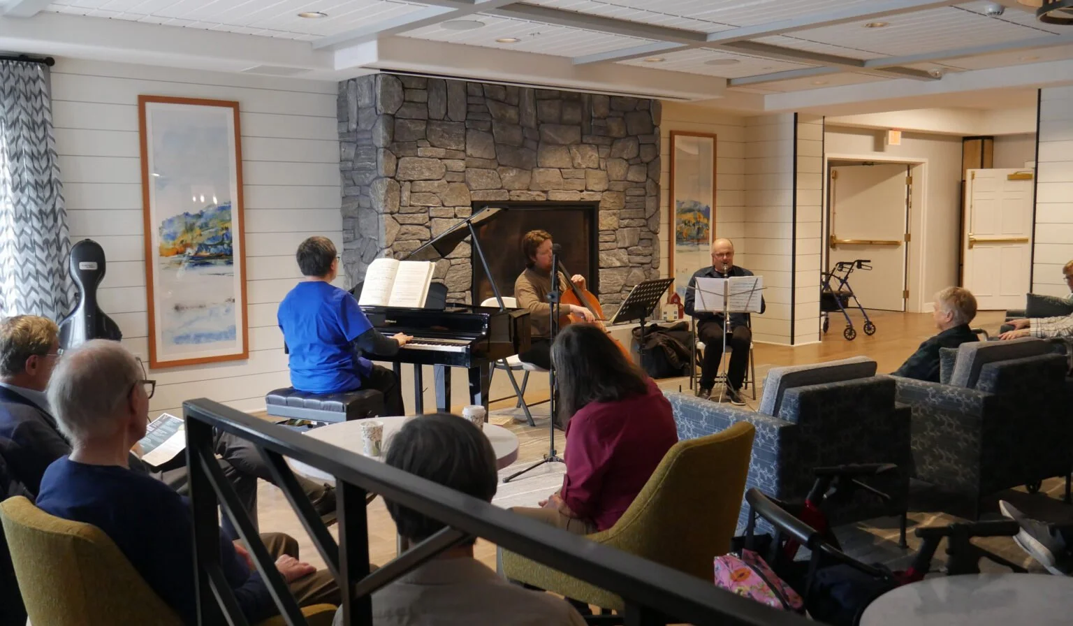 A live musical performance in a cozy room with a stone fireplace, a piano, a violinist, a pianist, and an audience seated in armchairs and chairs.