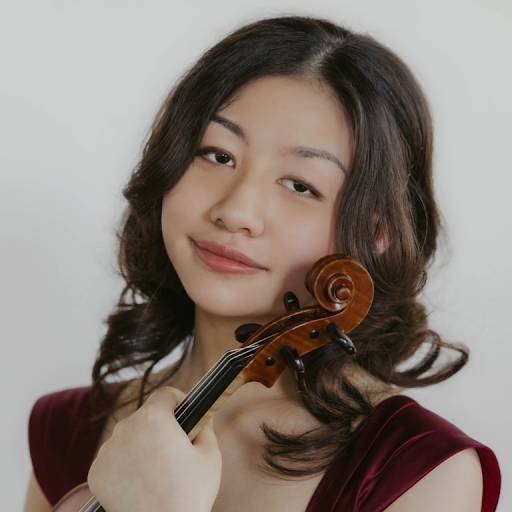 A young woman with wavy brown hair holding a violin close to her face, looking at the camera with a slight smile, wearing a dark red top.