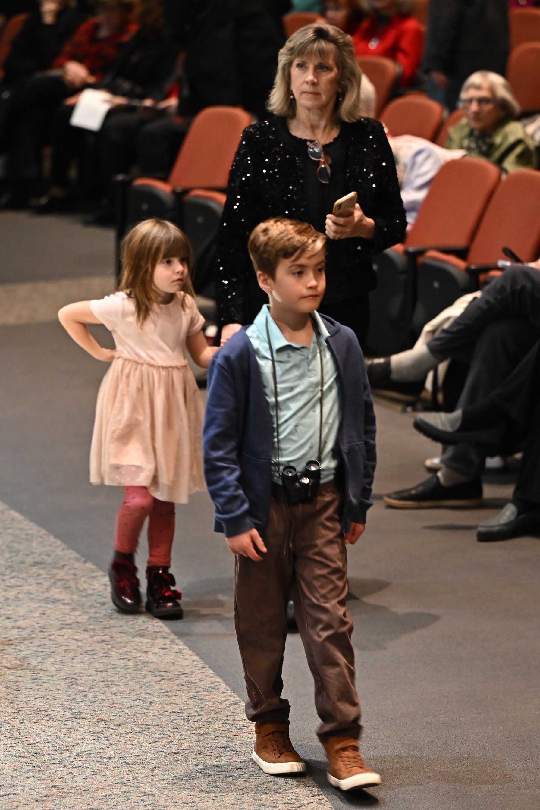 A woman with blonde hair wearing a black sequined jacket holds a phone while standing in an auditorium. A young girl with blonde hair in a pink dress with tights and shiny boots stands behind her, and a young boy with brown hair, a blue jacket, and binoculars around his neck walks in front of her. Other people are seated in rows of orange seats in the background.