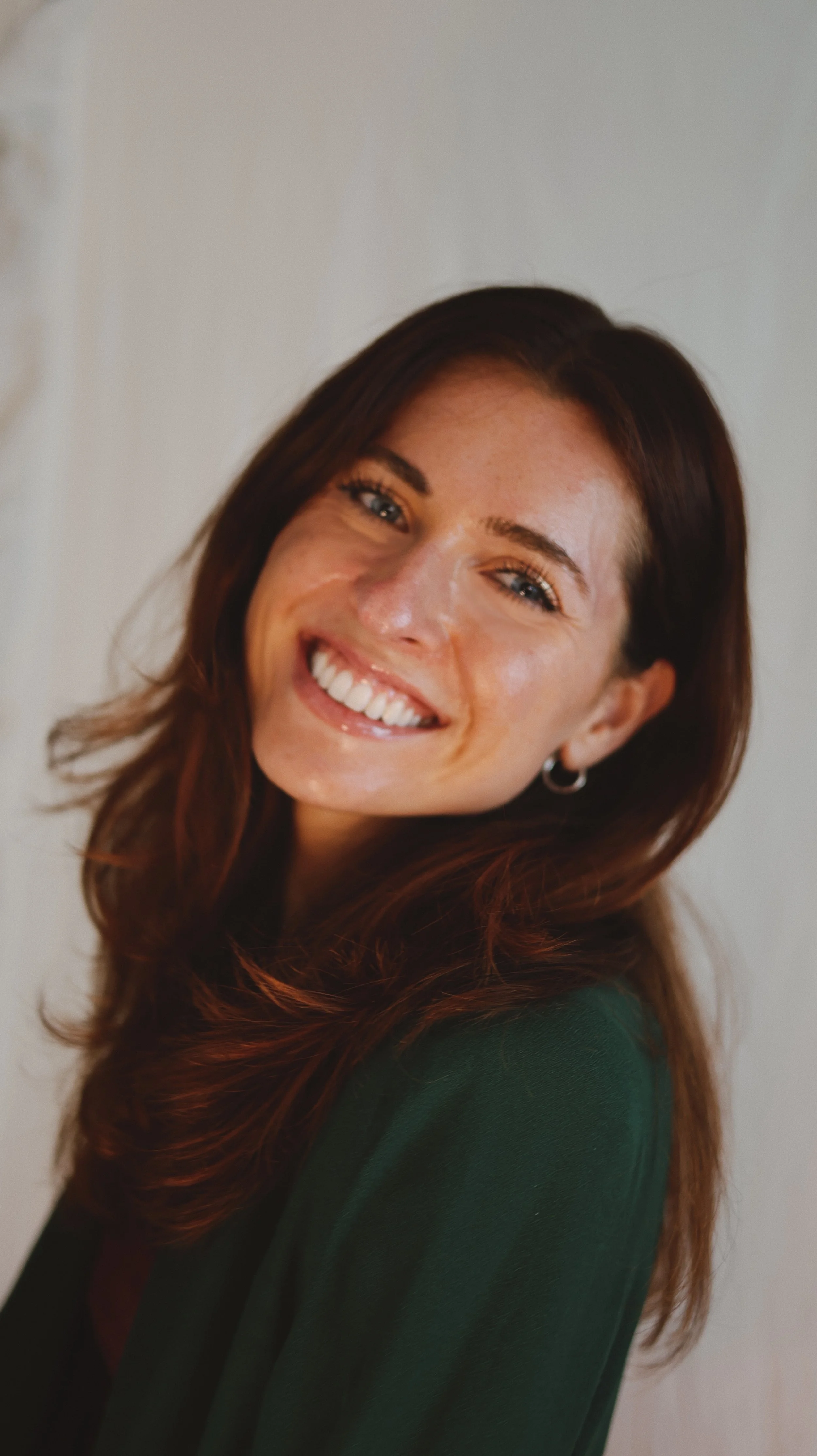 A woman with long, wavy auburn hair and blue eyes smiling, wearing a dark green top, standing against a plain background.