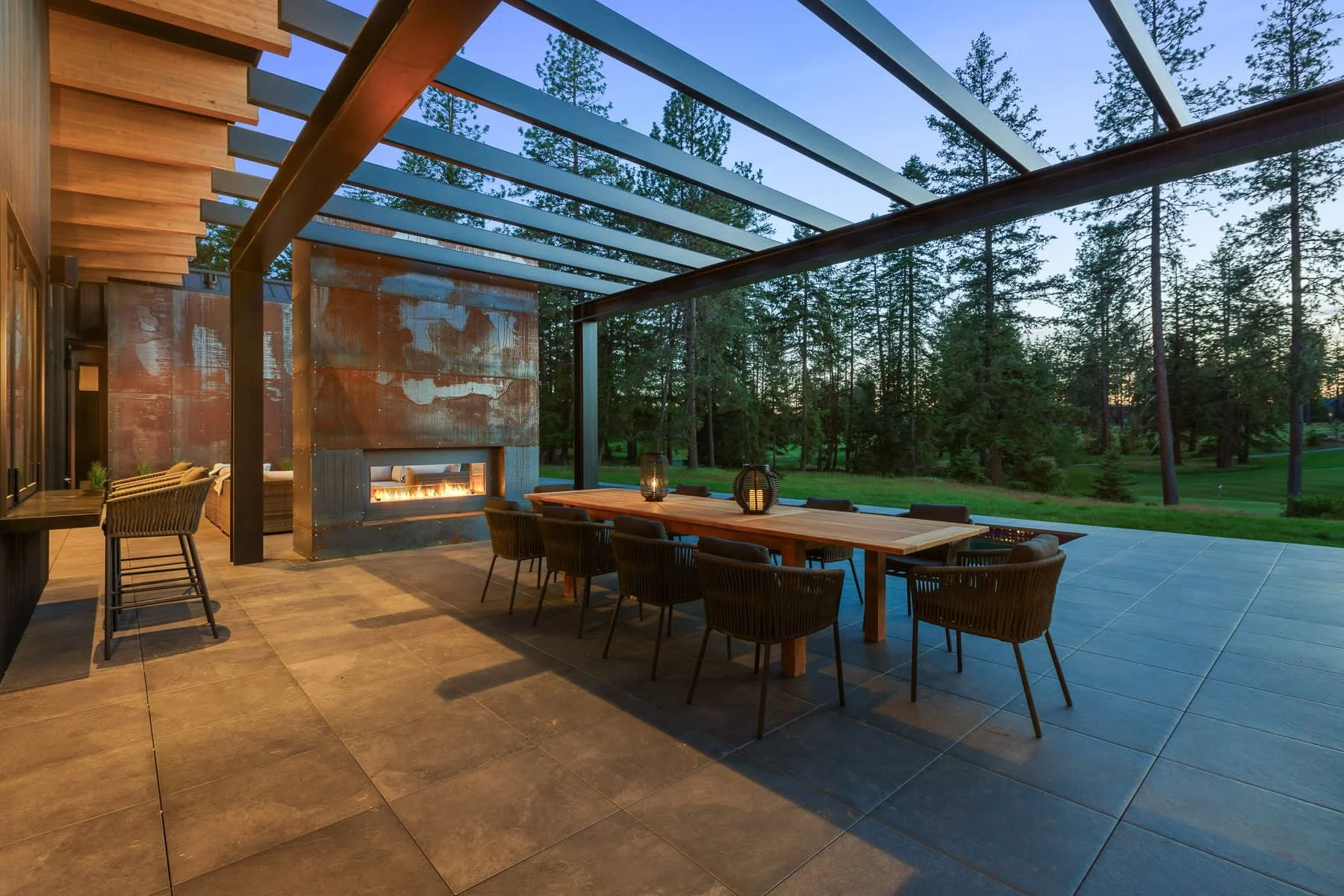 Outdoor patio with a large dining table and eight chairs, a modern fireplace, and a sitting area with a sofa. The patio is enclosed by a pergola with a view of trees and a grassy area at dusk.
