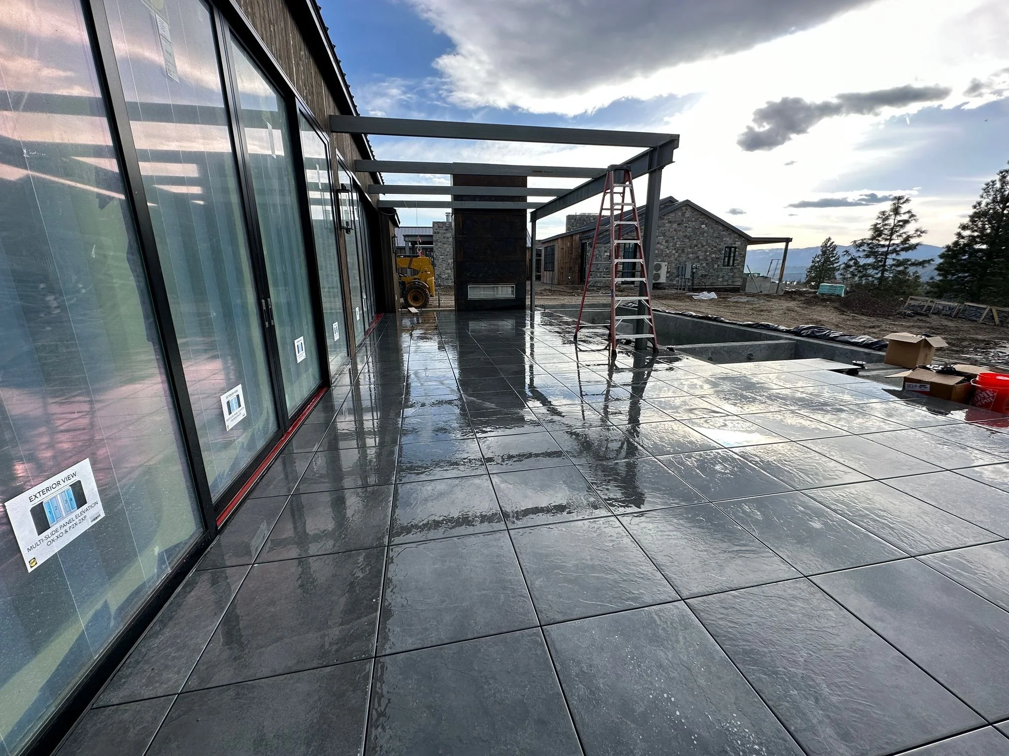 Construction site with glass doors, wet tiled floor, ladders, construction equipment, and nearby houses with mountains in the background under a partly cloudy sky.