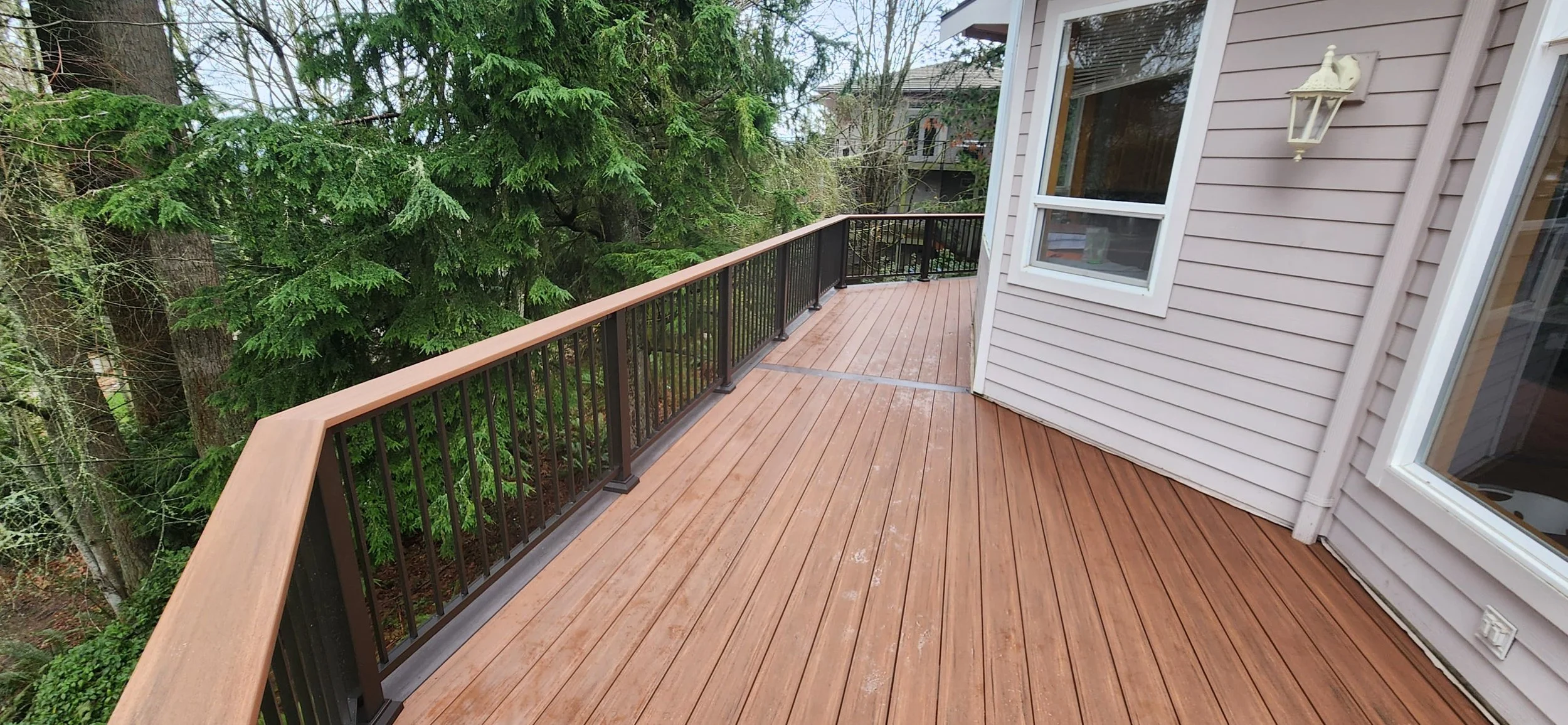 A wooden deck attached to a house with a black metal railing, overlooking green trees and neighboring houses in the background.