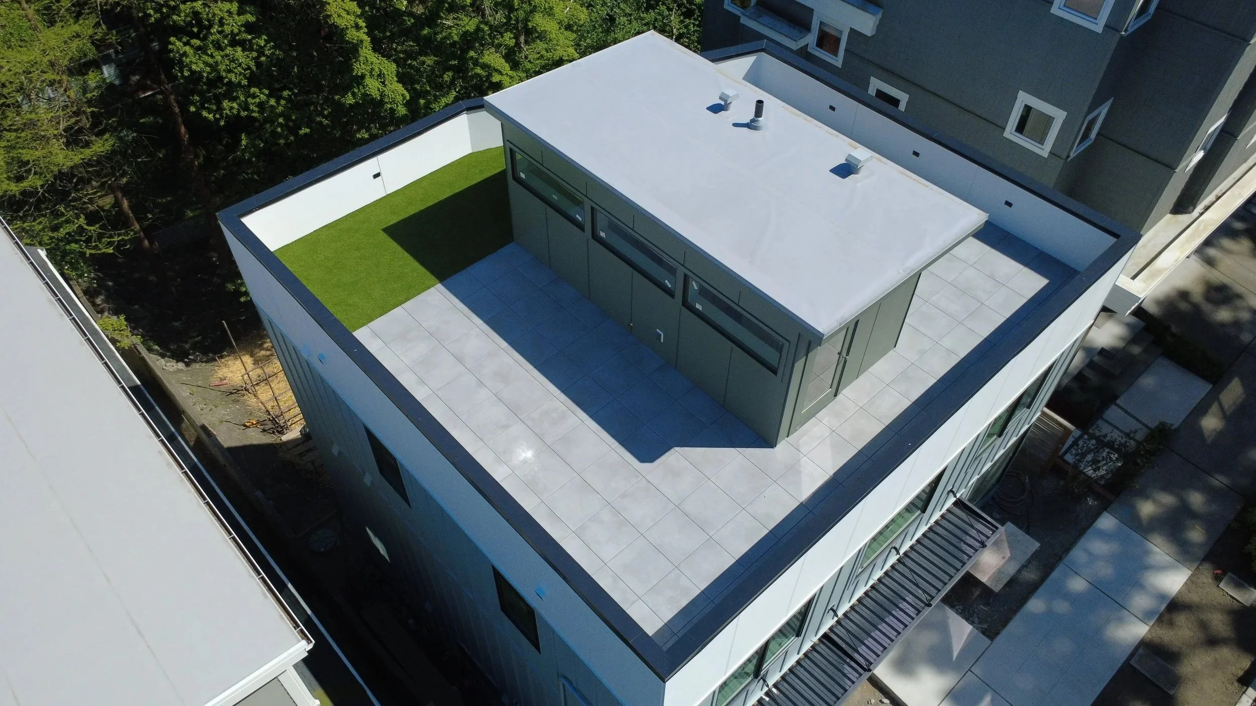 An aerial view of a modern building rooftop with a white and gray exterior, green artificial turf area, and surrounding trees.