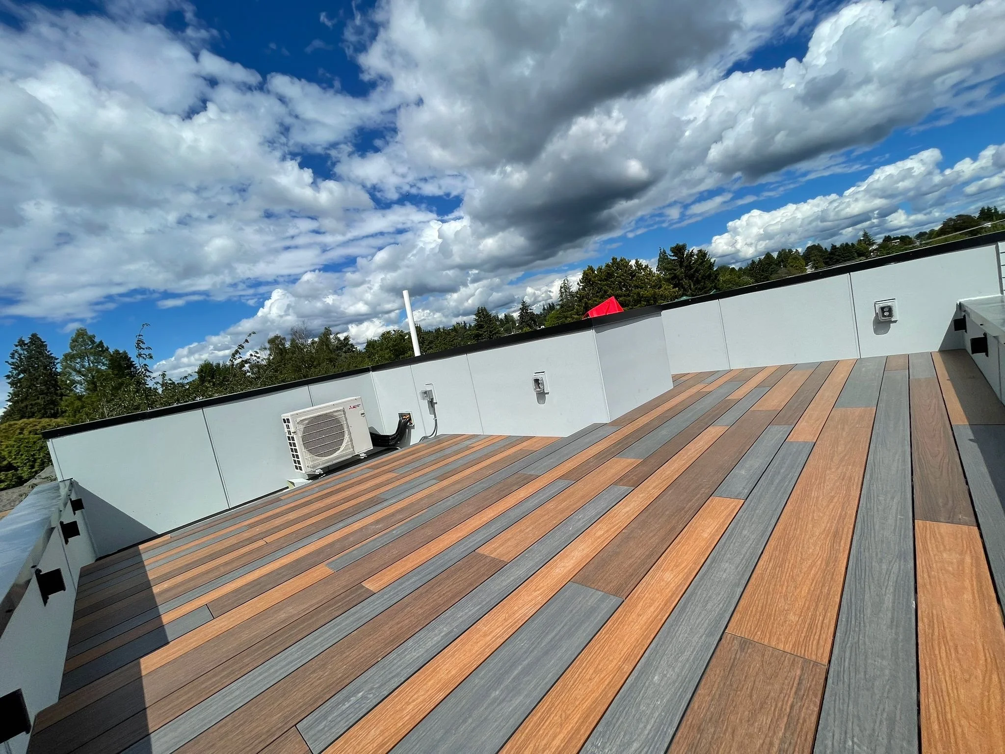 View of a rooftop deck with wood plank flooring, a white wall with a small air conditioning unit, and a cloudy blue sky overhead with trees in the background.