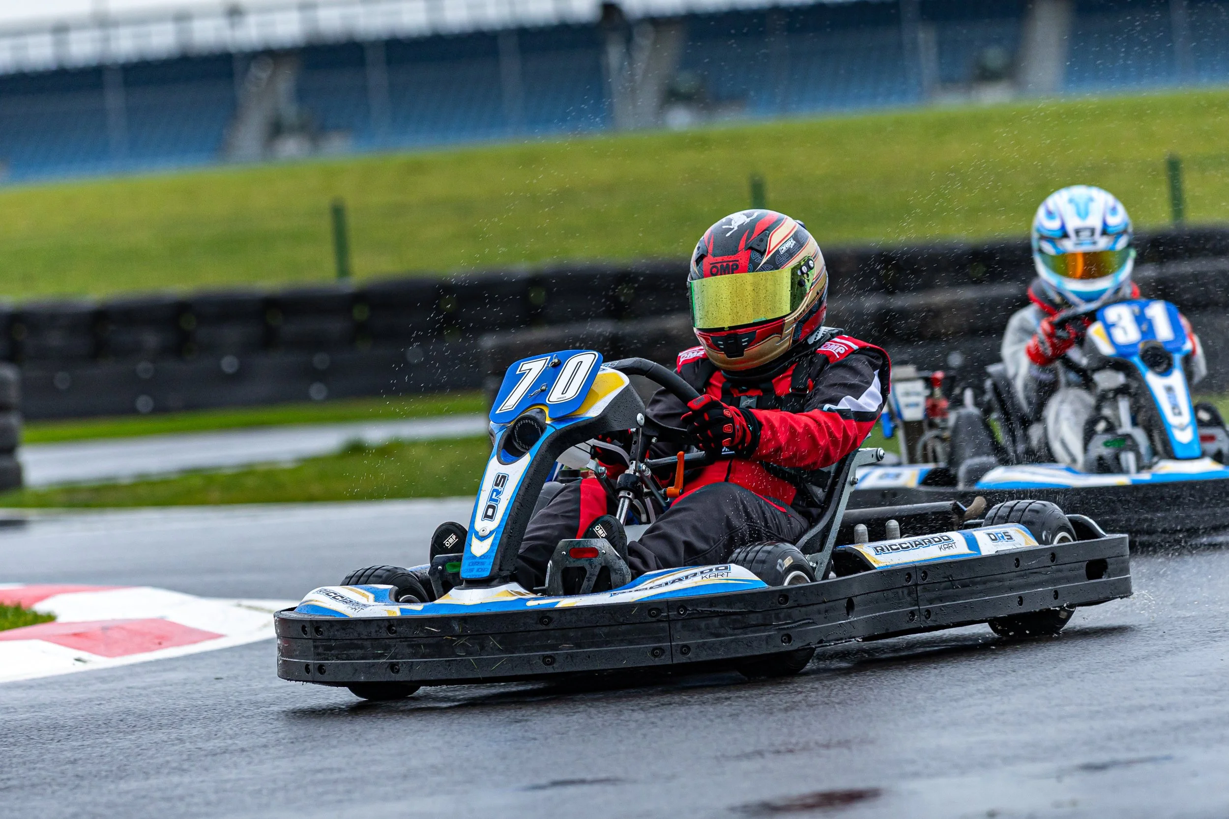 Two individuals racing go-karts on a wet track, wearing helmets and racing gear. The track has visible water spray, indicating rainy conditions.