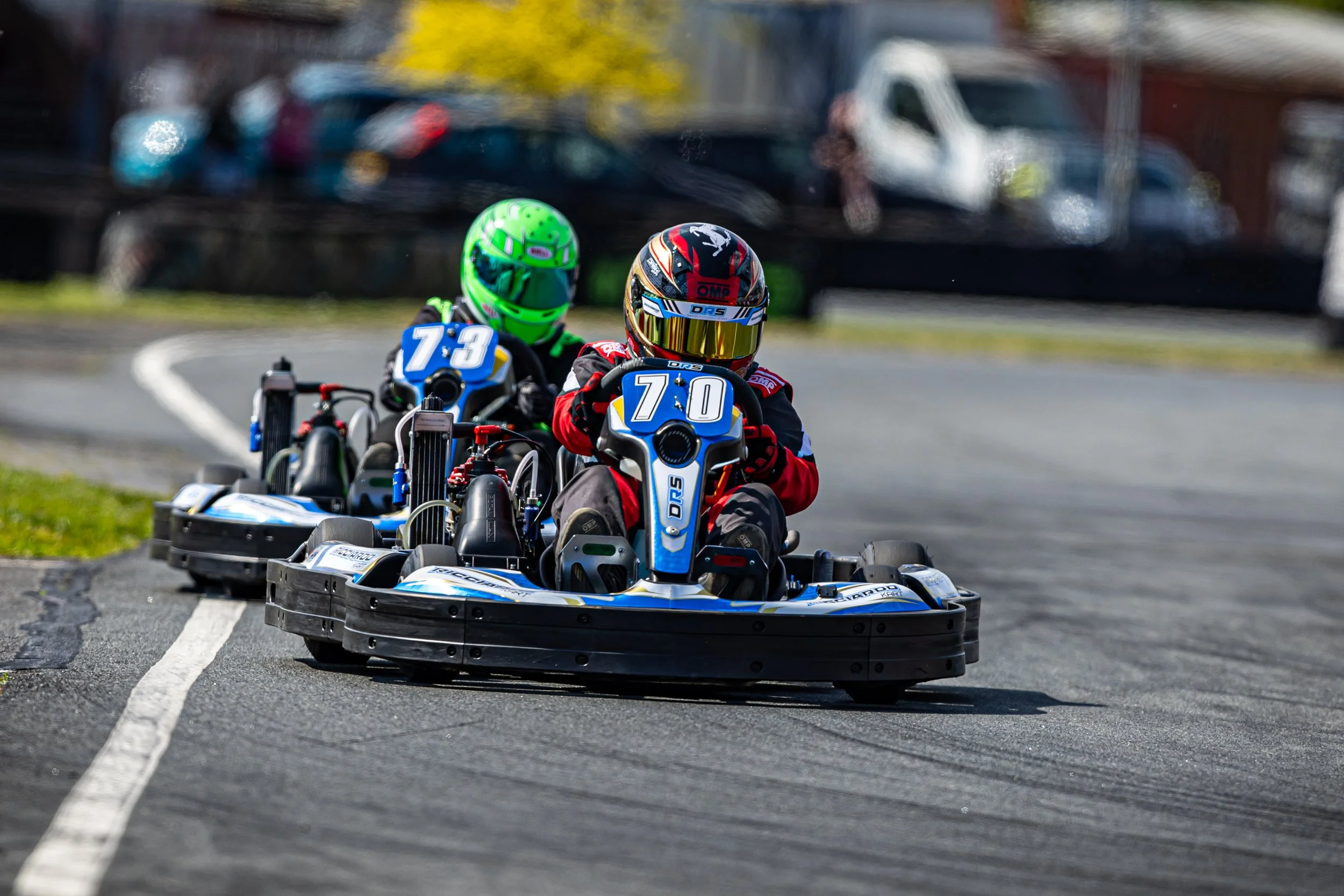 Two individuals racing go-karts on a track, both wearing helmets and racing suits, with blurred cars in the background.