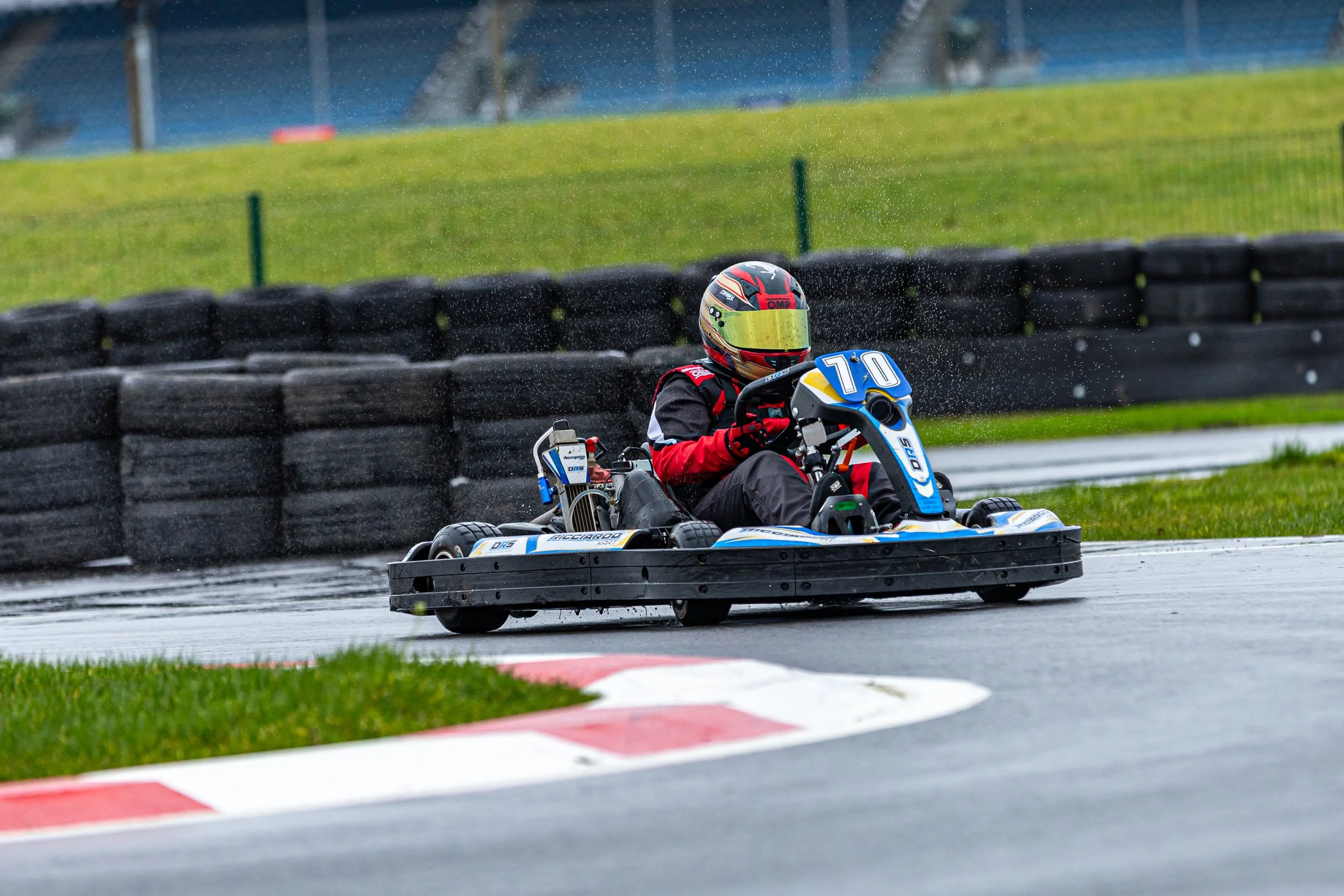 A go-kart driver wearing a black and red racing suit and a helmet, racing on a wet track, with water spray coming off the tires, and a row of tires acting as a barrier along the track.